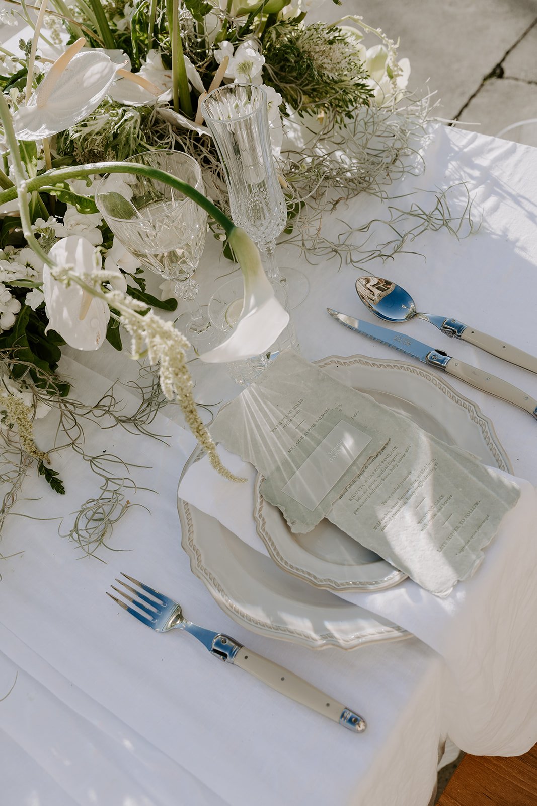 Table setting with white china plates, crystal glasses, silverware, surrounded by white flowers and greenery. There is a script paper or menu placed on the plate, and a salad server and fork are set on a white tablecloth.