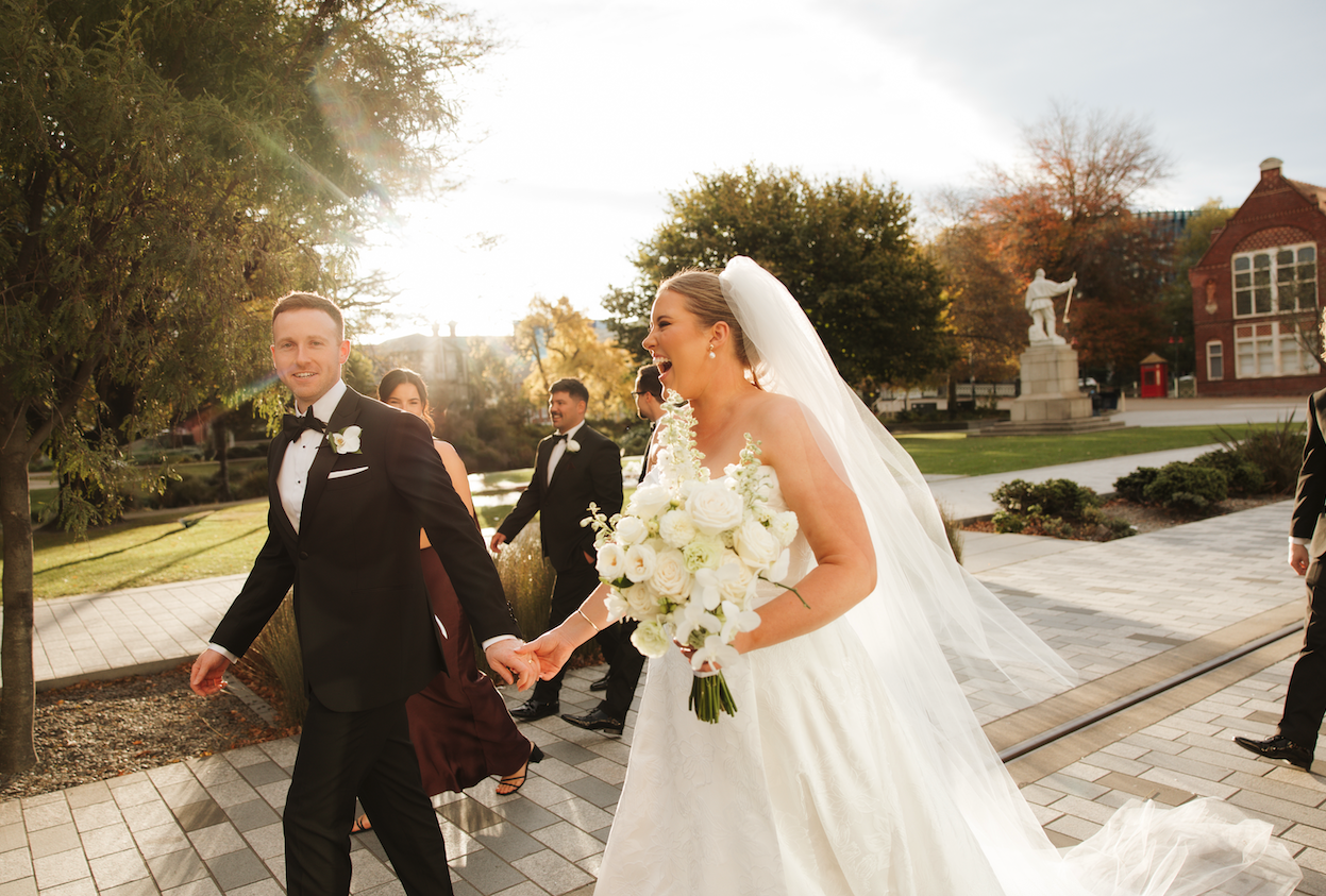 Newlyweds holding hands and smiling outside a brick building in golden sunlight.