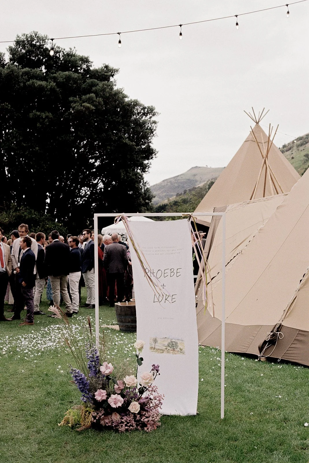 Wedding welcome sign with pastel flowers beside a tipi and festoon lights.