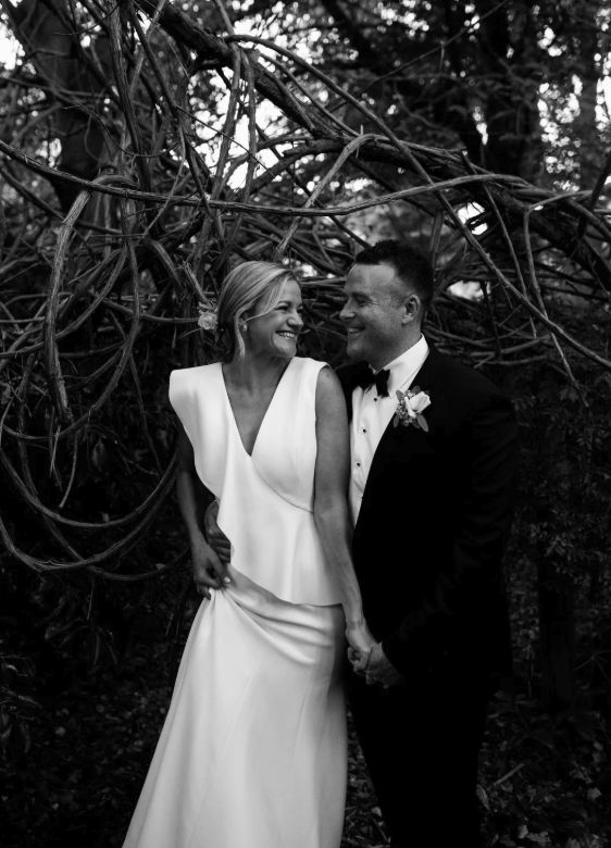 A wedding couple in formal attire, smiling at each other, holding hands outdoors in front of tree branches.
