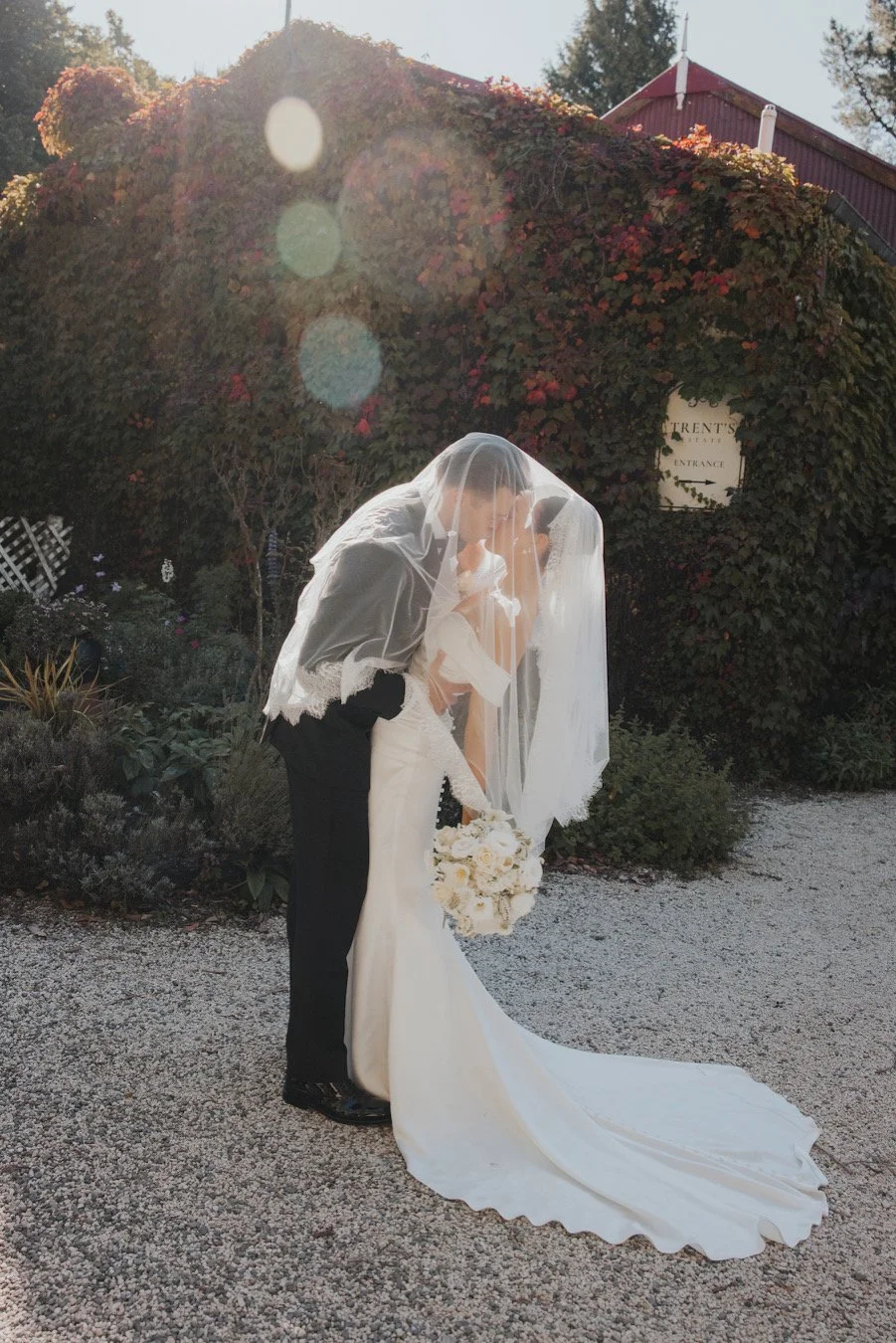 Bride and groom share a kiss under a veil outdoors at sunset, with a garden and arched vine-covered wall in the background.