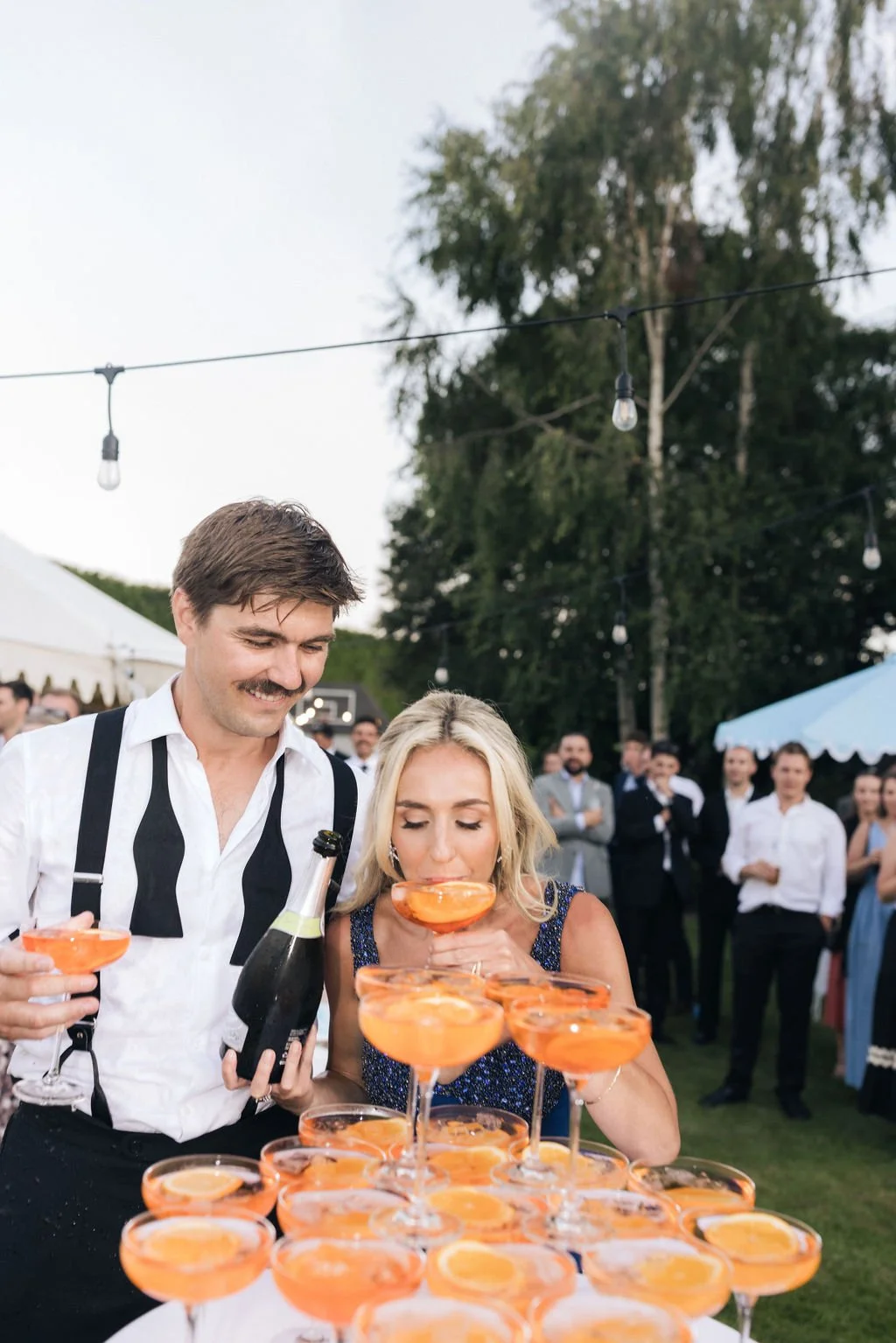 A man and a woman at an outdoor event, with the woman drinking from a cocktail glass filled with an orange beverage and slices of orange, while the man holds a bottle of champagne. There are numerous similar cocktails on the table in front of them, and people dressed in formal attire are gathered in the background.