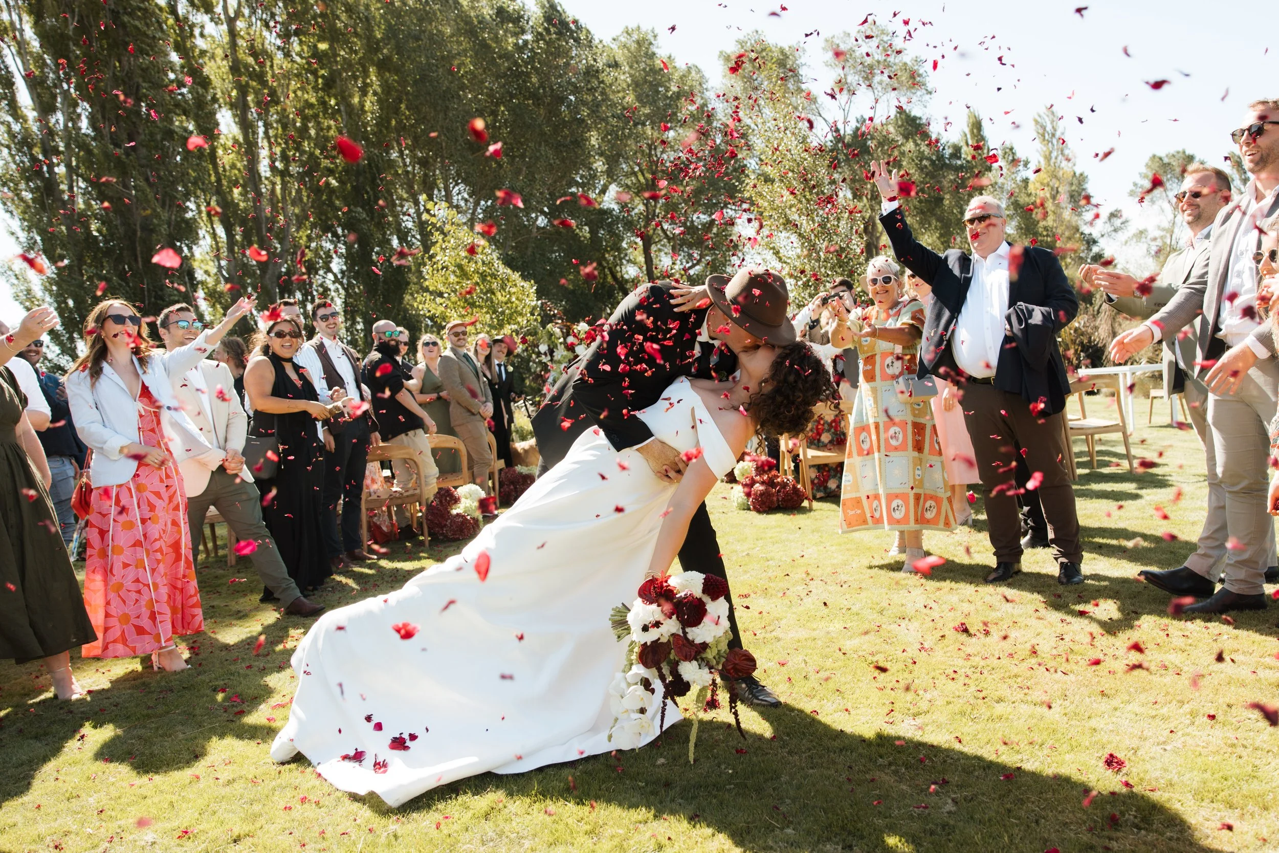 A wedding ceremony outdoors with a bride and groom kissing, surrounded by friends and family. Rose petals are thrown in the air, and the wedding party is smiling and enjoying the celebration in a sunny, green park.