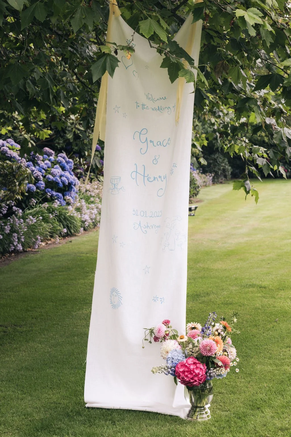 White fabric wedding welcome sign hanging beneath greenery with flowers at its base.