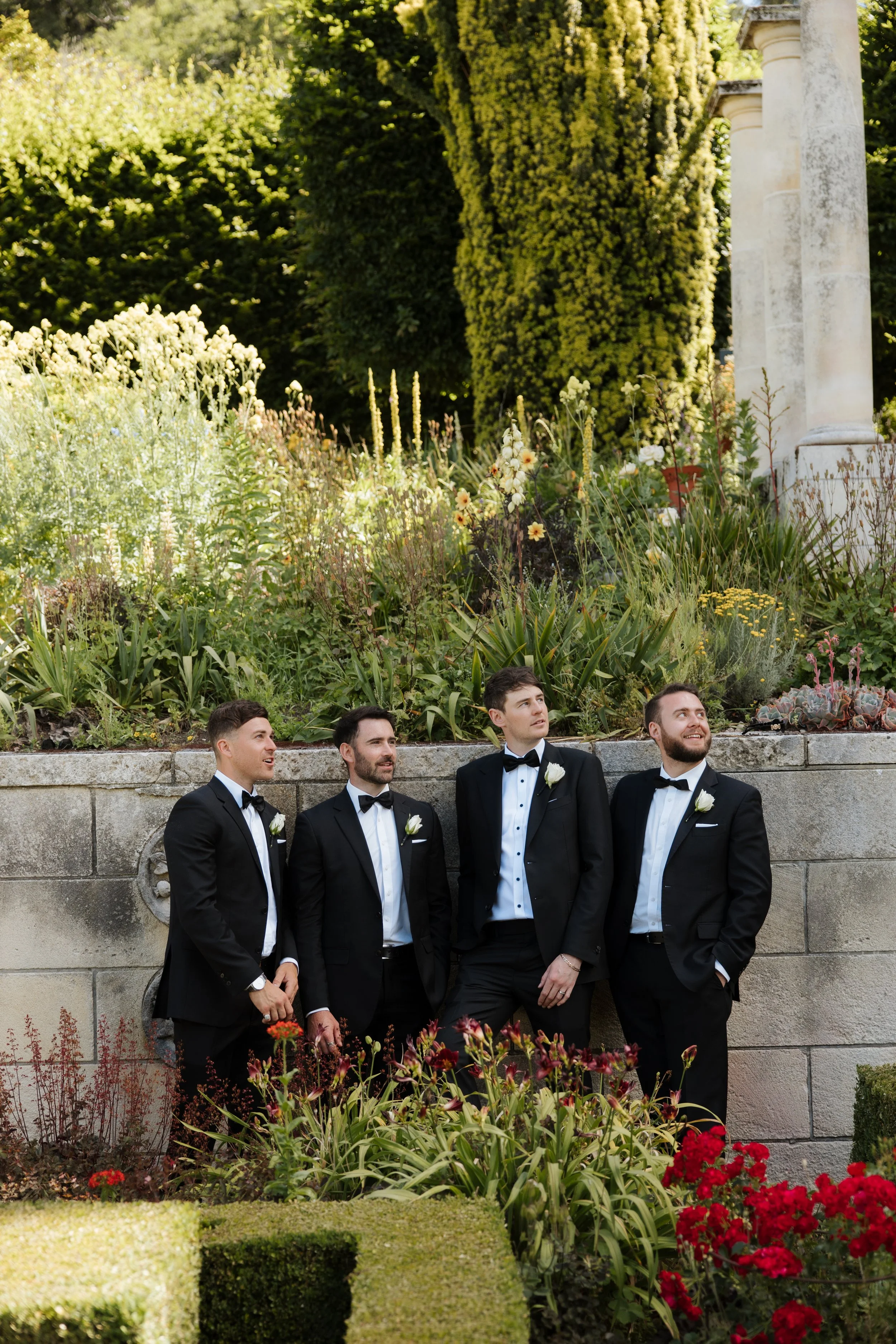 Four men in tuxedos and bow ties standing together outdoors near a stone wall with lush greenery and flowers in the background.