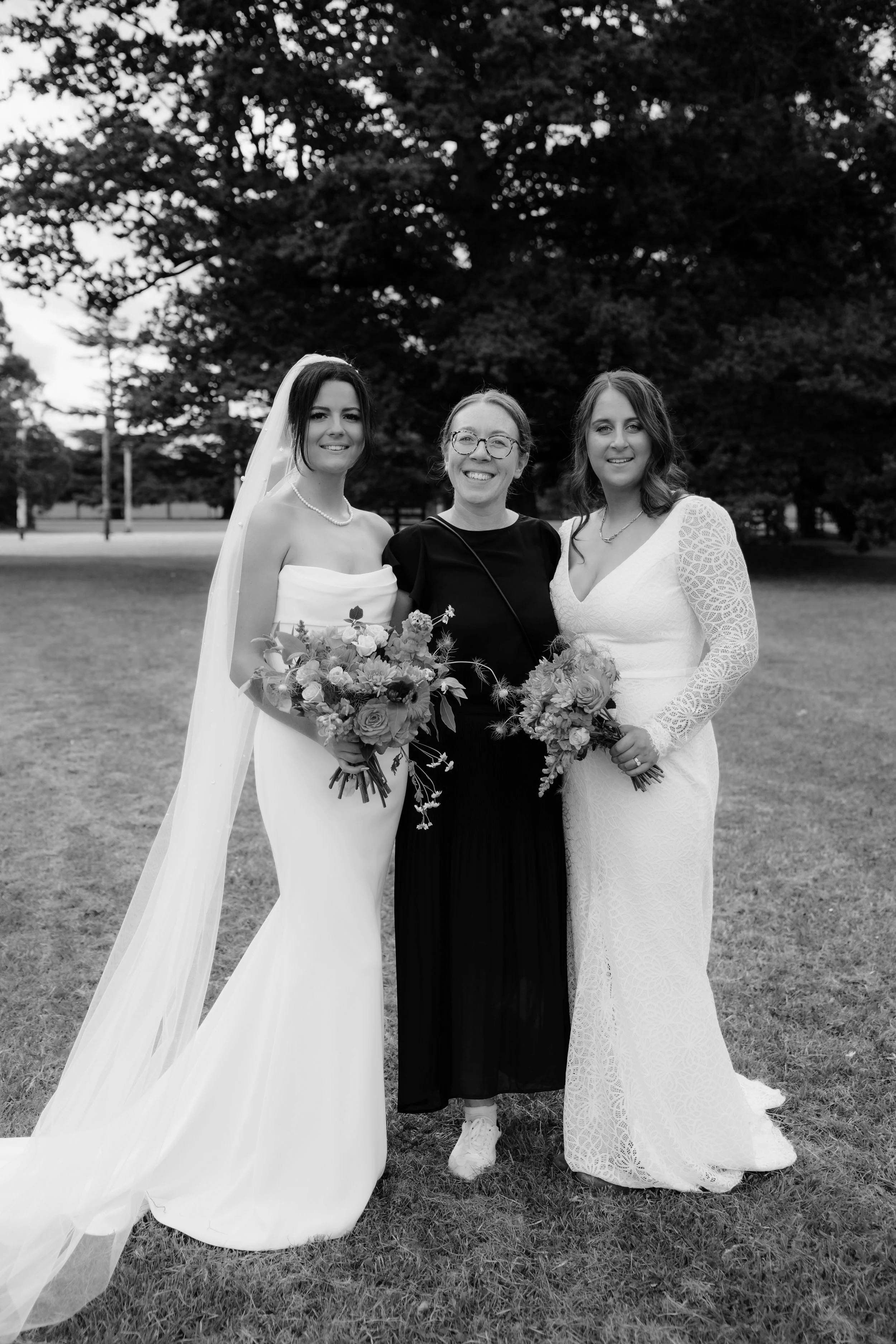 Three women standing outdoors, two in wedding dresses holding bouquets, one in black clothing, all smiling for a photo with trees in the background.