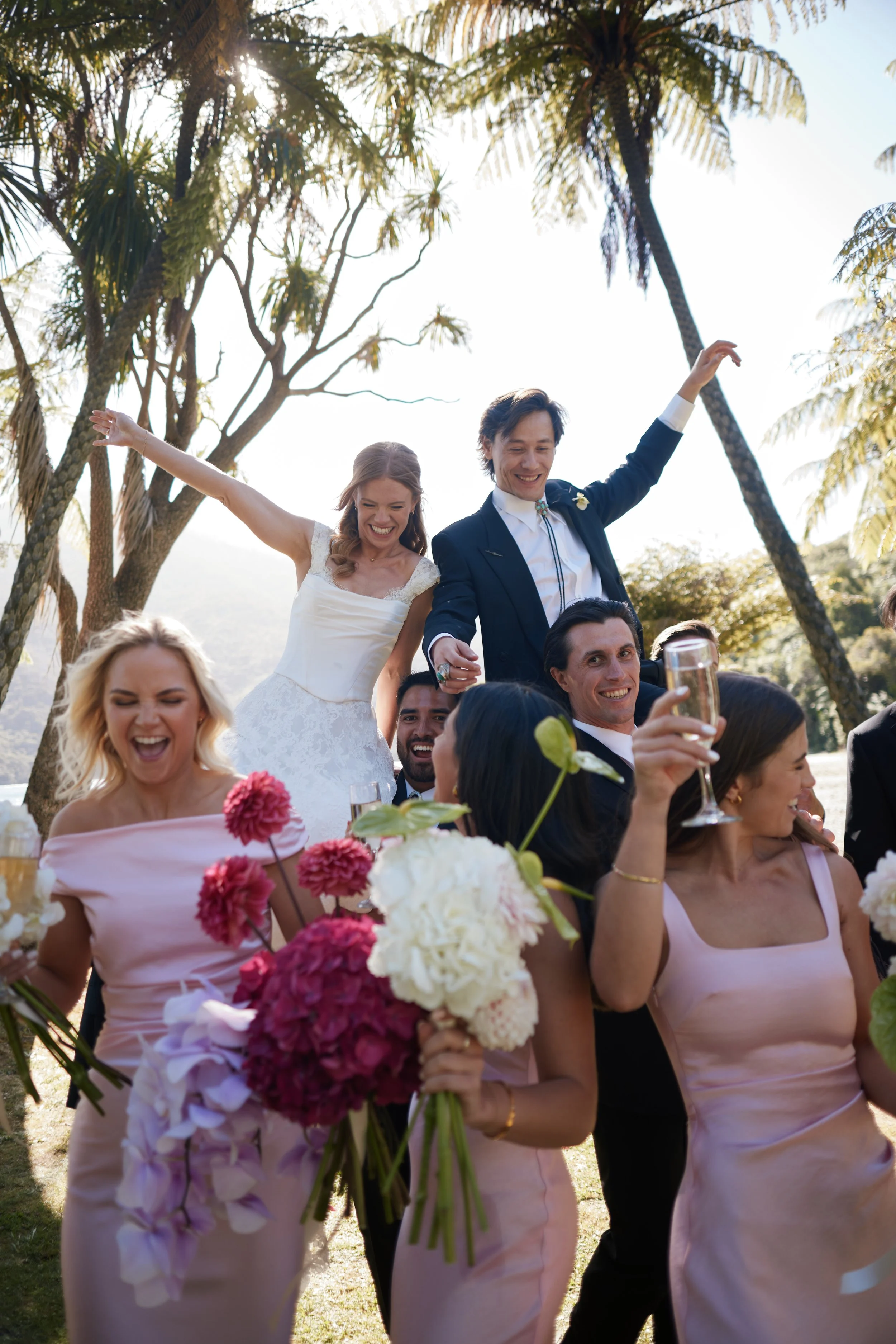 A wedding celebration outdoors with a bride and groom standing on a group of people, holding hands and smiling, surrounded by palm trees and bright sunlight, with pink and white flowers in the foreground.