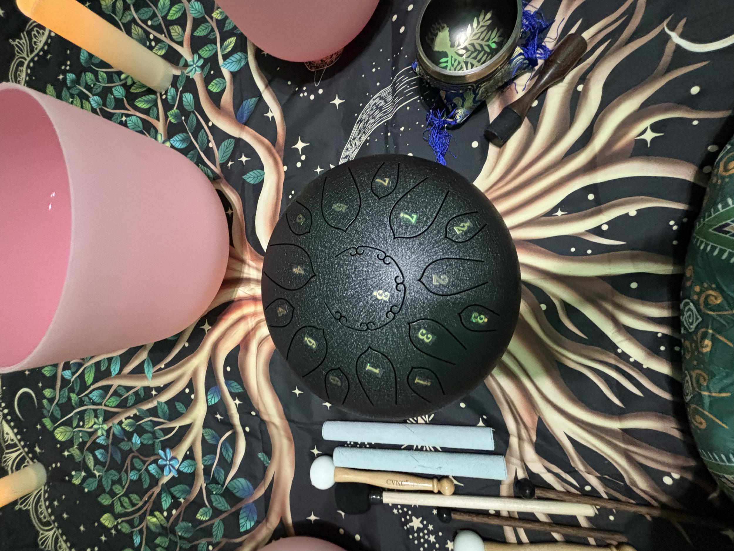 A collection of musical instruments including a black handpan, several mallets, a pink drum, a candle, a jar, a rolled-up paper, and balloons, all on a decorative tablecloth with a tree and star pattern.