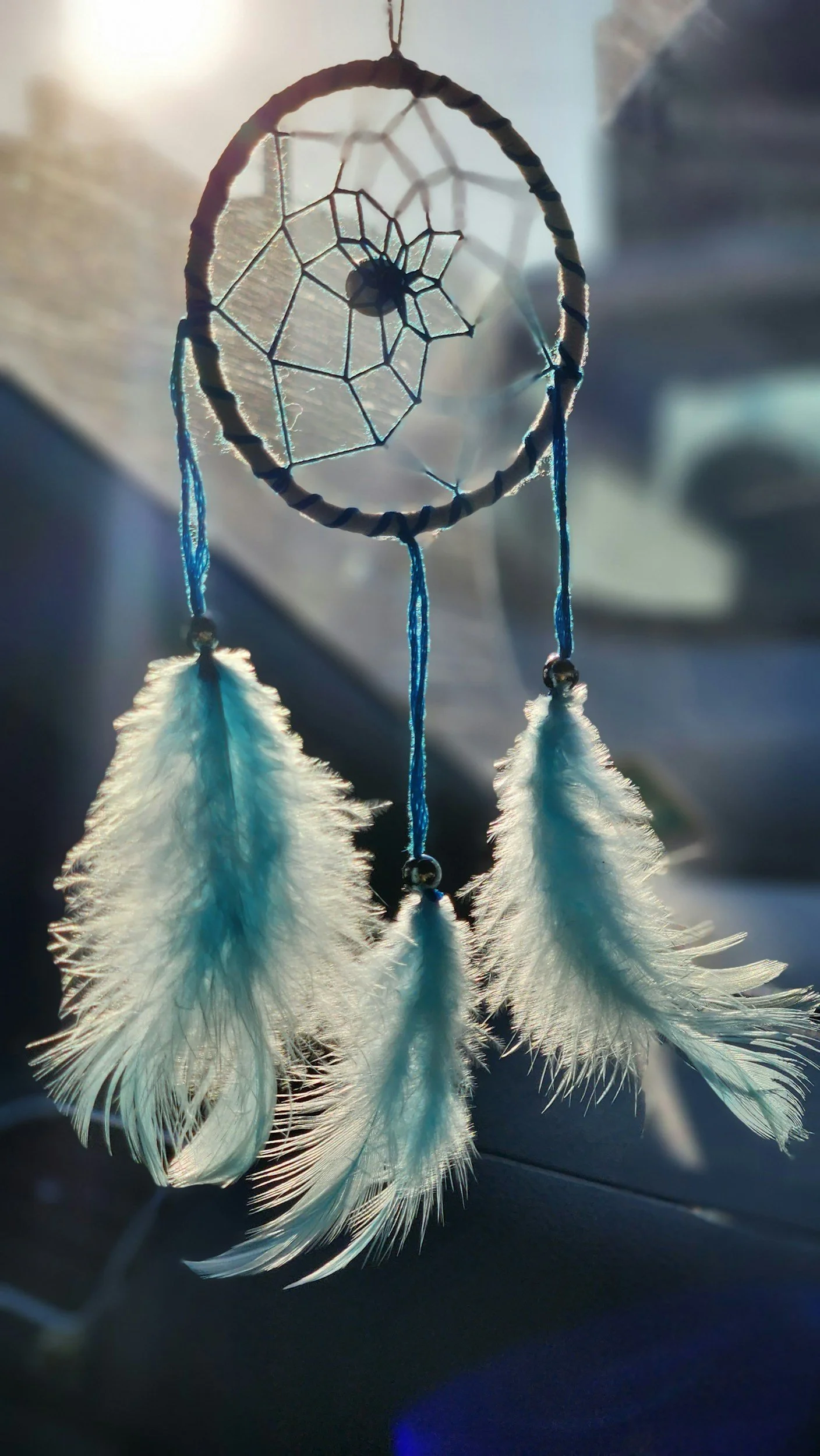 Close-up of a dreamcatcher with a small spiderweb center, three white feathers hanging down, backlit by sunlight.