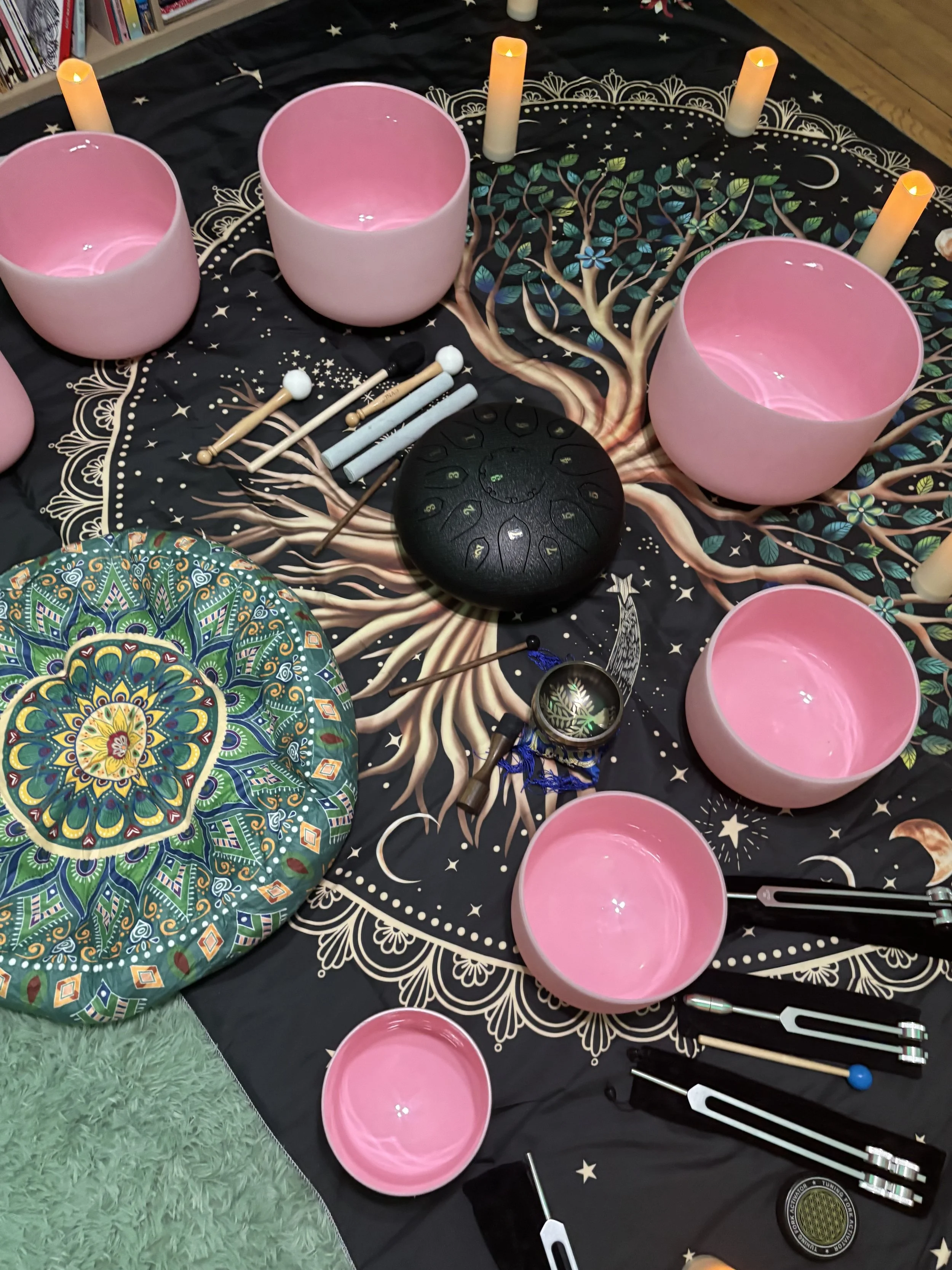 A set of pink crystal singing bowls, a black meditation chime, and various mallets on a black cloth with a mystical tree design, surrounded by candles and decorative items.