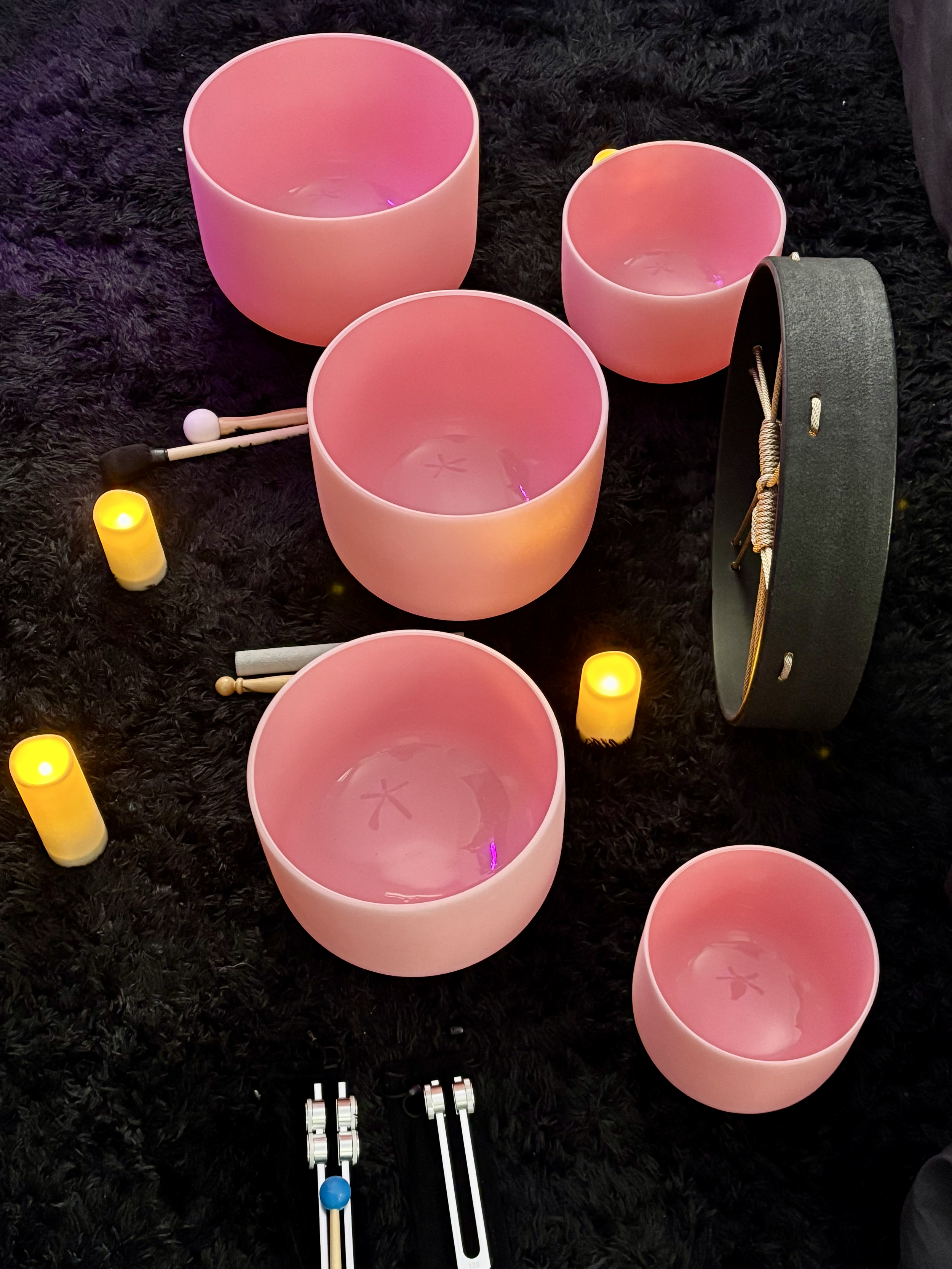 Five pink crystal singing bowls arranged on a black fuzzy surface, illuminated by small yellow LED candles, with a black-and-gold shaker, a pair of mallets, and a tambourine nearby.