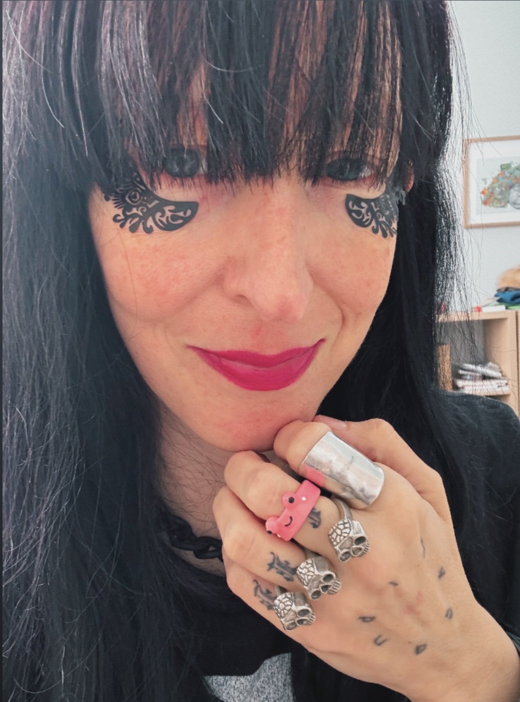 A woman with long black hair and bangs, wearing bright pink lipstick, is smiling slightly. She has dark eye tattoos under her eyes and is wearing multiple rings, including a pink toy ring and skull-shaped rings. The background shows a bookshelf and s