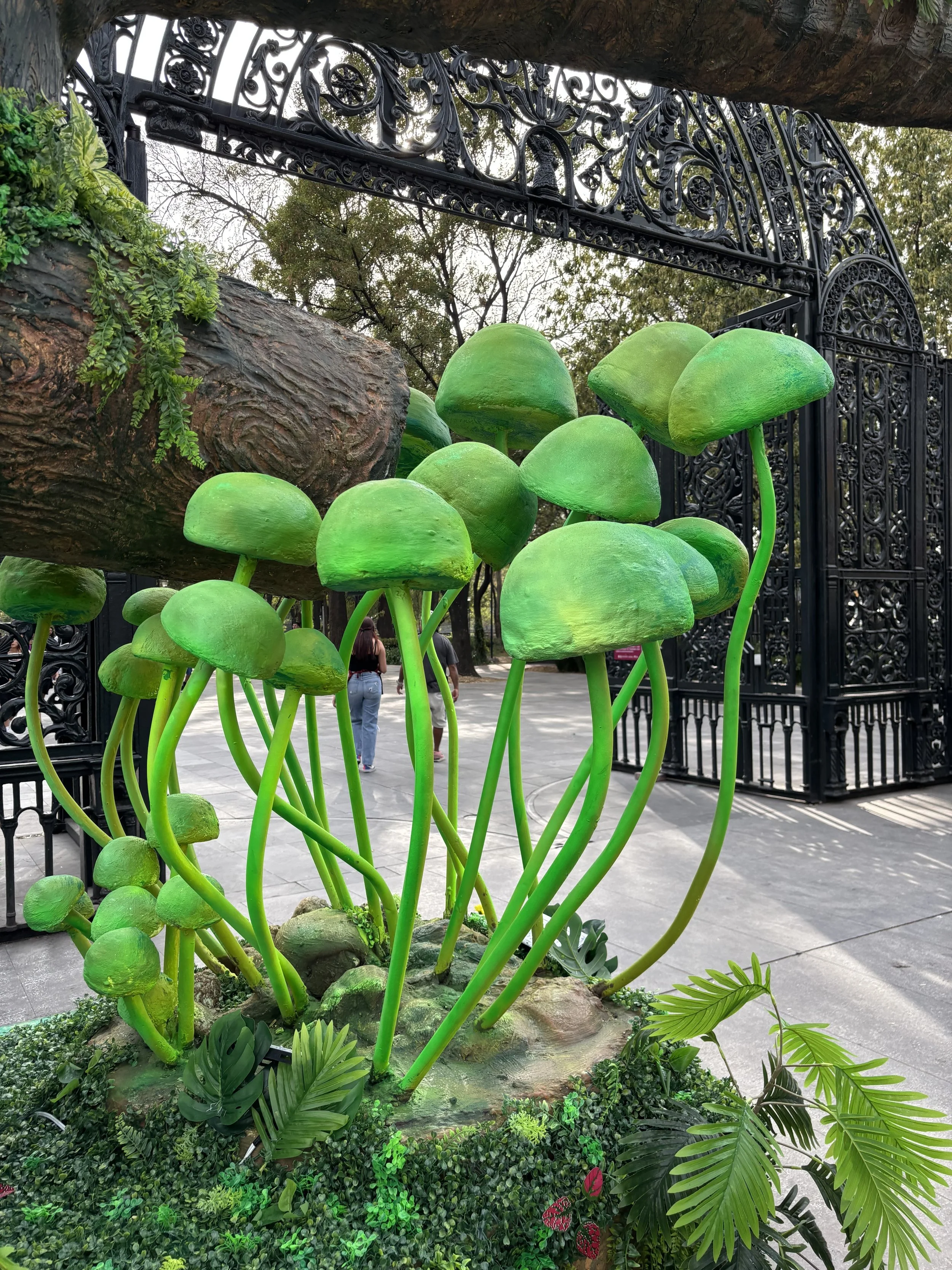 Bright green mushroom sculptures with curved stems and rounded caps, set in a garden area with greenery and an ornate black metal gate in the background.