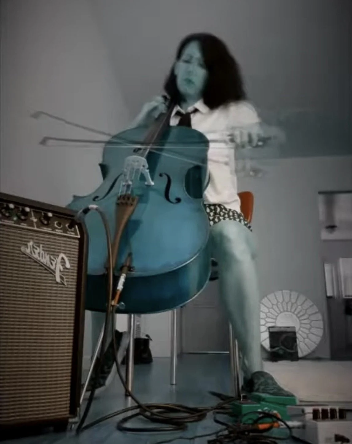 A person with long dark hair, wearing a white shirt and black tie, is sitting on a stool playing a teal-colored acoustic guitar, with a vintage amplifier nearby, in a room with gray walls.