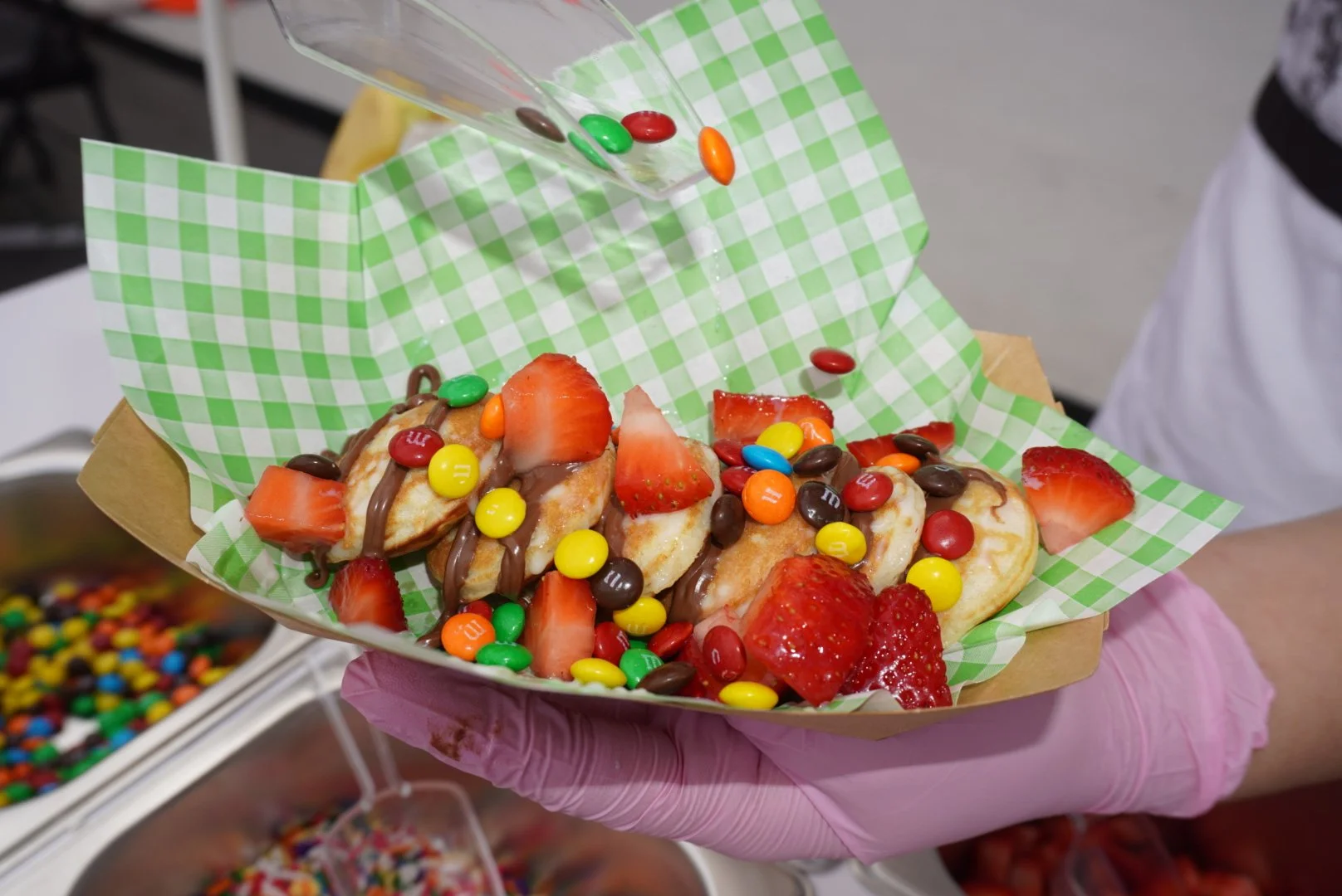A person holding a tray with mini waffles topped with sliced strawberries, chocolate drizzle, and M&M candies, with more M&Ms in a container nearby.