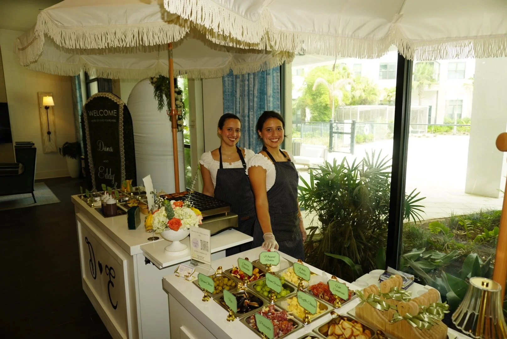Two women wearing aprons standing behind a table with assorted small food dishes and a floral arrangement, in a bright indoor space with large windows and greenery outside.