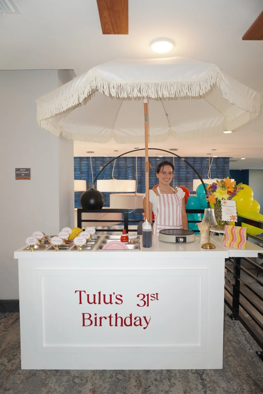A birthday setup for Tulu's 31st birthday featuring a woman standing behind a white counter with a large umbrella shading her, colorful balloons, a bouquet of flowers, and birthday decorations.