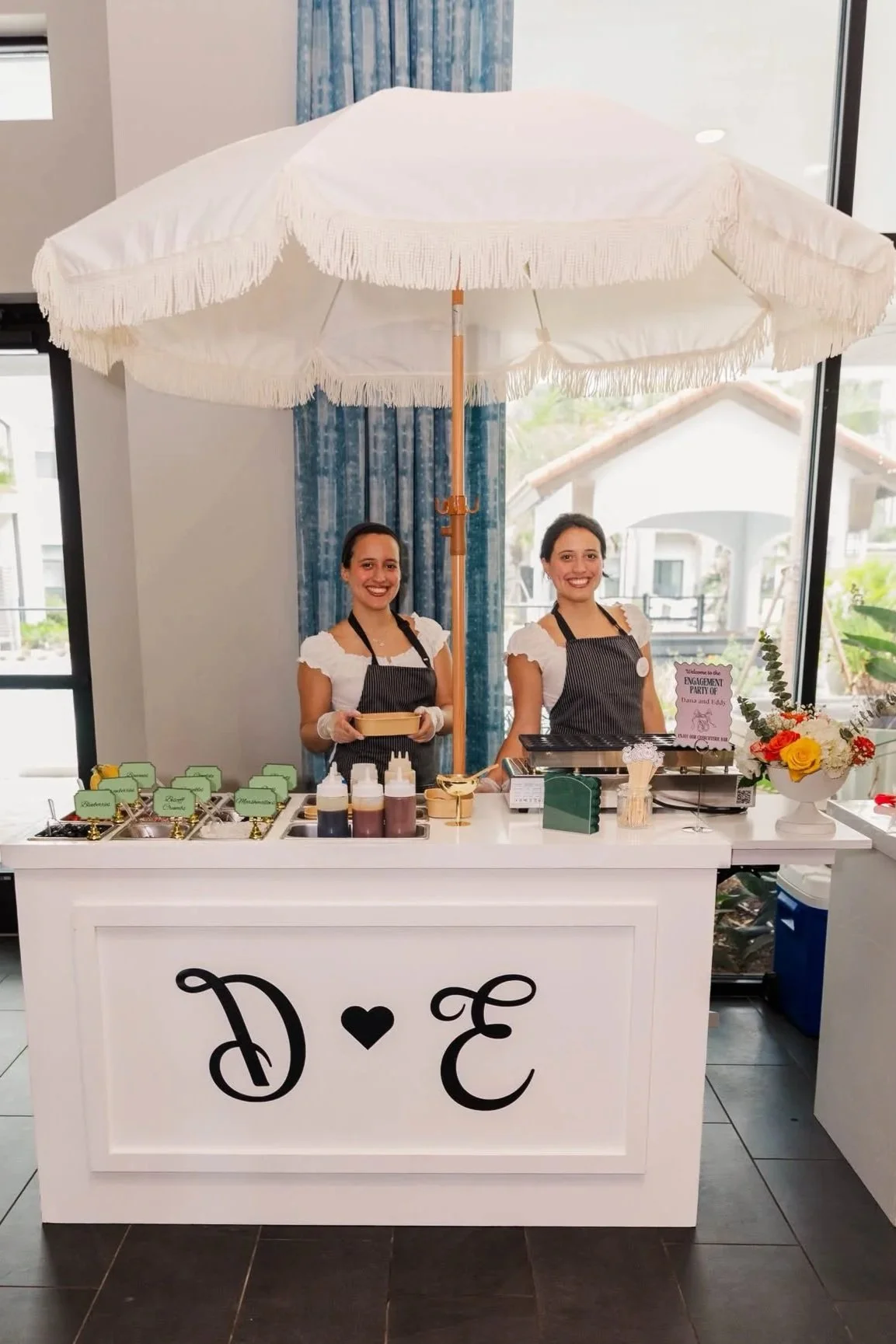 Two women standing behind a dessert or food stand decorated with a large white umbrella, flowers, and a sign that reads 'D + E'. The women are smiling and wearing black aprons.