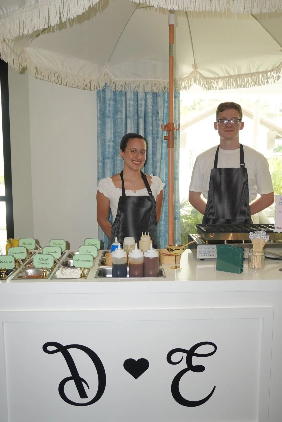 Two people standing behind a dessert stand with various toppings and sauces, under a large white umbrella. The stand has a sign with the text 'D E' and a heart symbol, and appears to be set up for a special event or celebration.