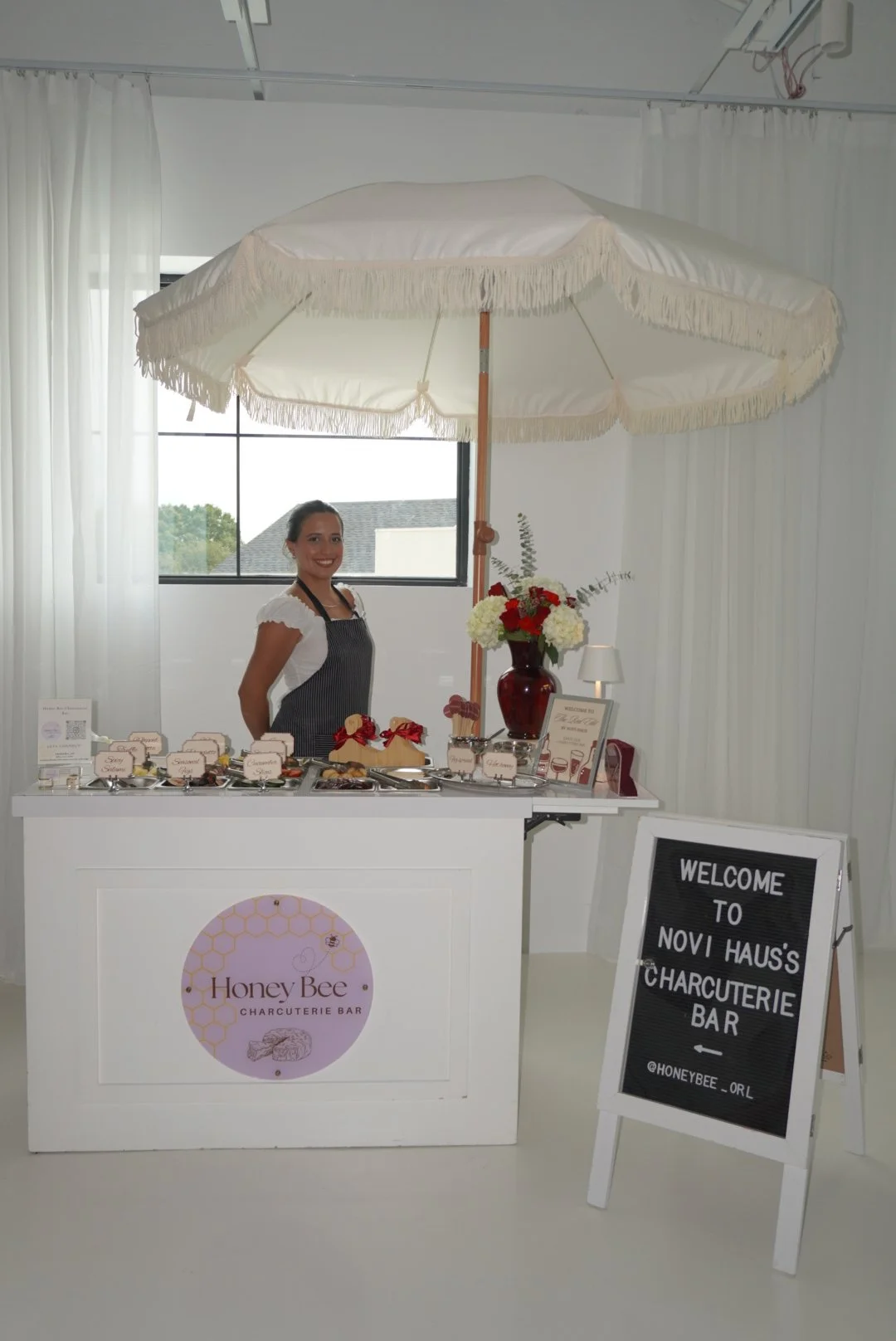A woman standing behind a white charcuterie bar with a sign that says "Honey Bee Charcuterie Bar." The bar has small plates of food, decorative signs, and a large floral arrangement. There is a large umbrella overhead and a chalkboard sign with white