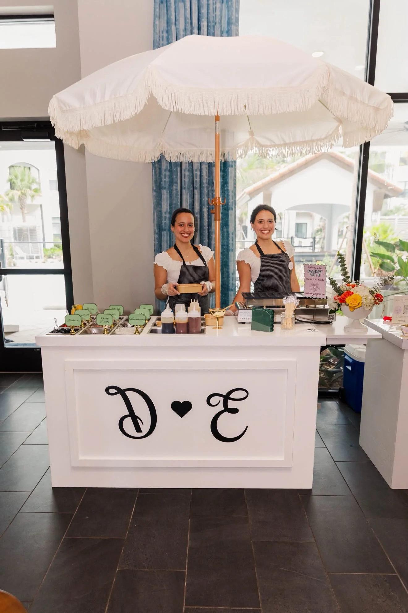 A dessert stand decorated with an umbrella and flowers, manned by two women in uniform, displaying jars of sauces and a sign for a bridal engagement party.