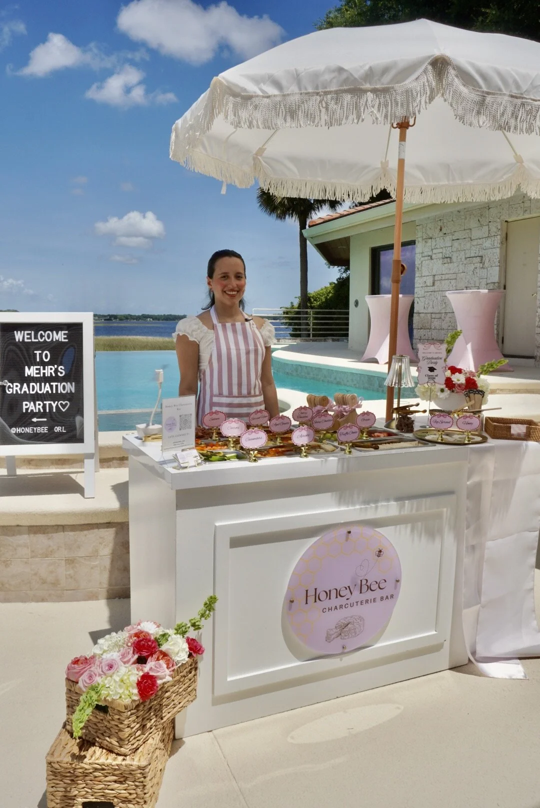 A woman standing behind a white charcuterie bar labeled 'Honey Bee Charcuterie Bar' at an outdoor poolside event, with tables of food, pink flowers, and an umbrella, under a blue sky.