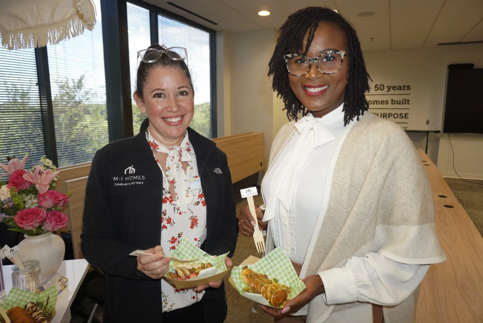 Two women standing indoors at an event, holding food in green checkered trays, smiling at the camera. One woman wears glasses, a black jacket, and a floral blouse, with a party badge. The other woman has glasses, a white bow-tie blouse, and a beige s
