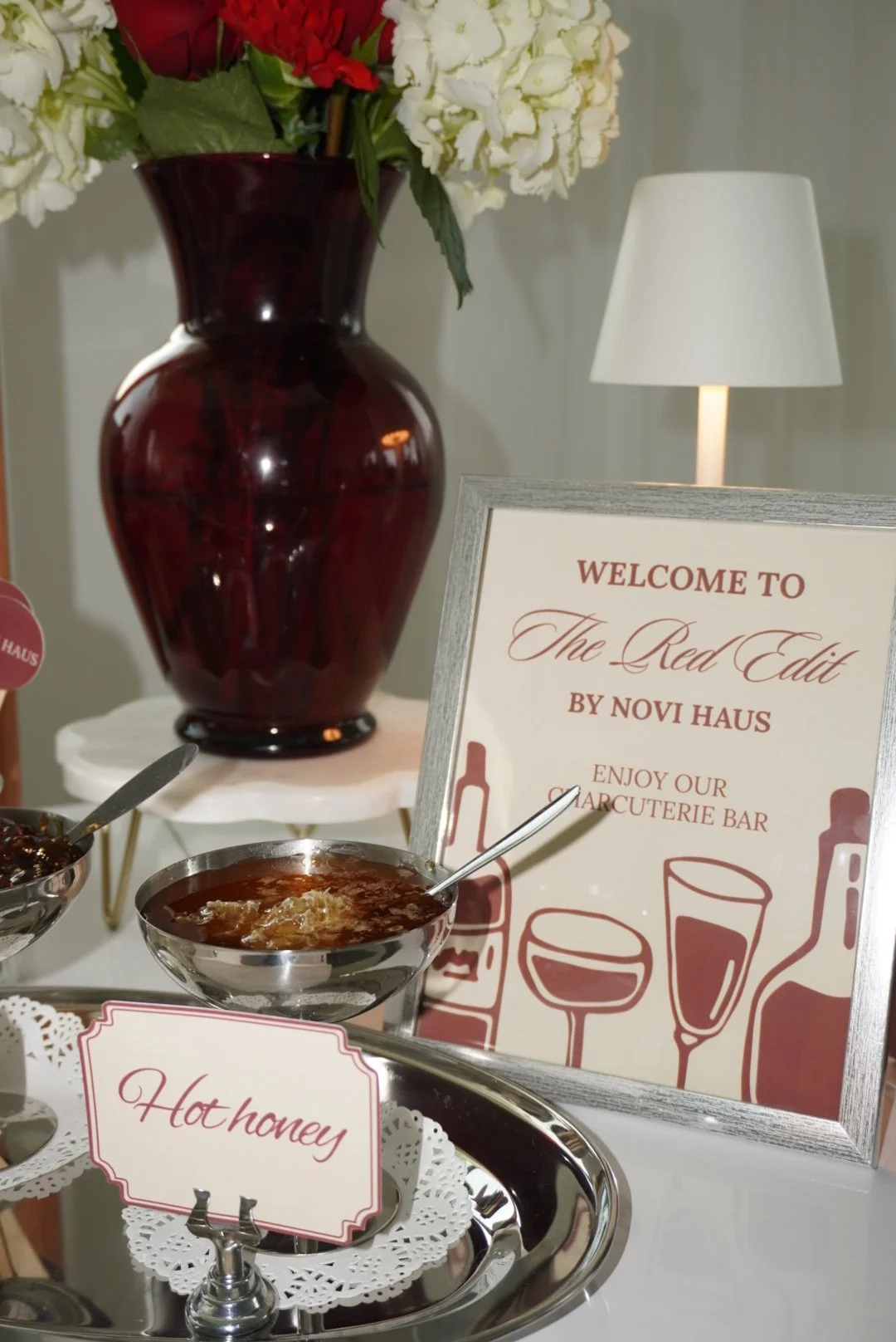 A floral arrangement in a dark red vase, a sign welcoming guests to The Red Edit by Novi Haus and inviting them to enjoy the marquetterie bar, small bowls with honey and jam, and a white lamp in the background.