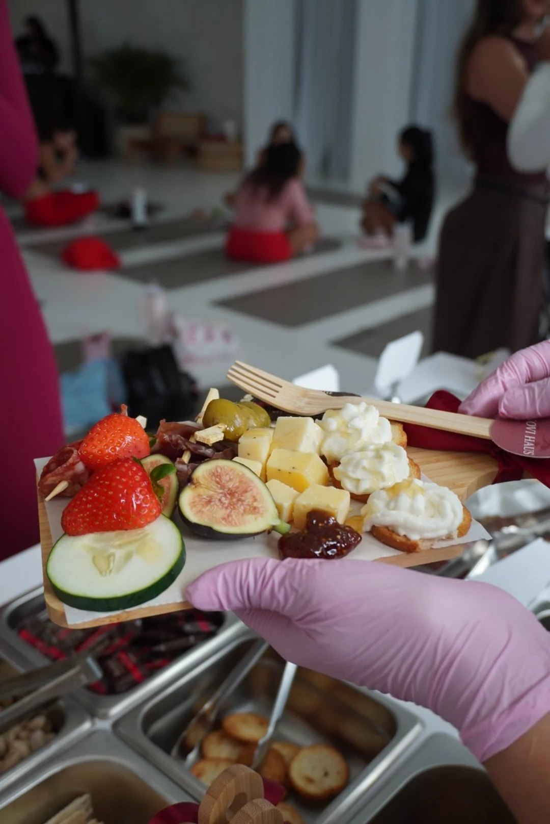 A tray of assorted cheeses, strawberries, figs, olives, and bread with whipped cream, on a buffet table in a room with people in the background.
