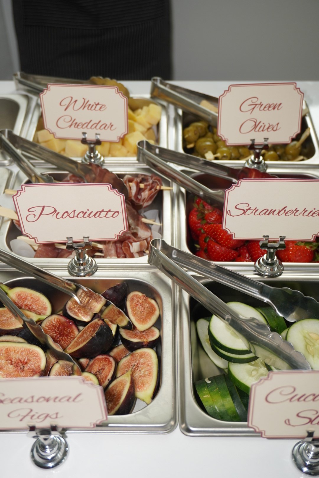 A buffet with various fresh fruits and vegetables including strawberries, figs, cucumbers, and green olives. There are also labels indicating different items such as white cheddar, prosciutto, strawberries, green olives, and figs.
