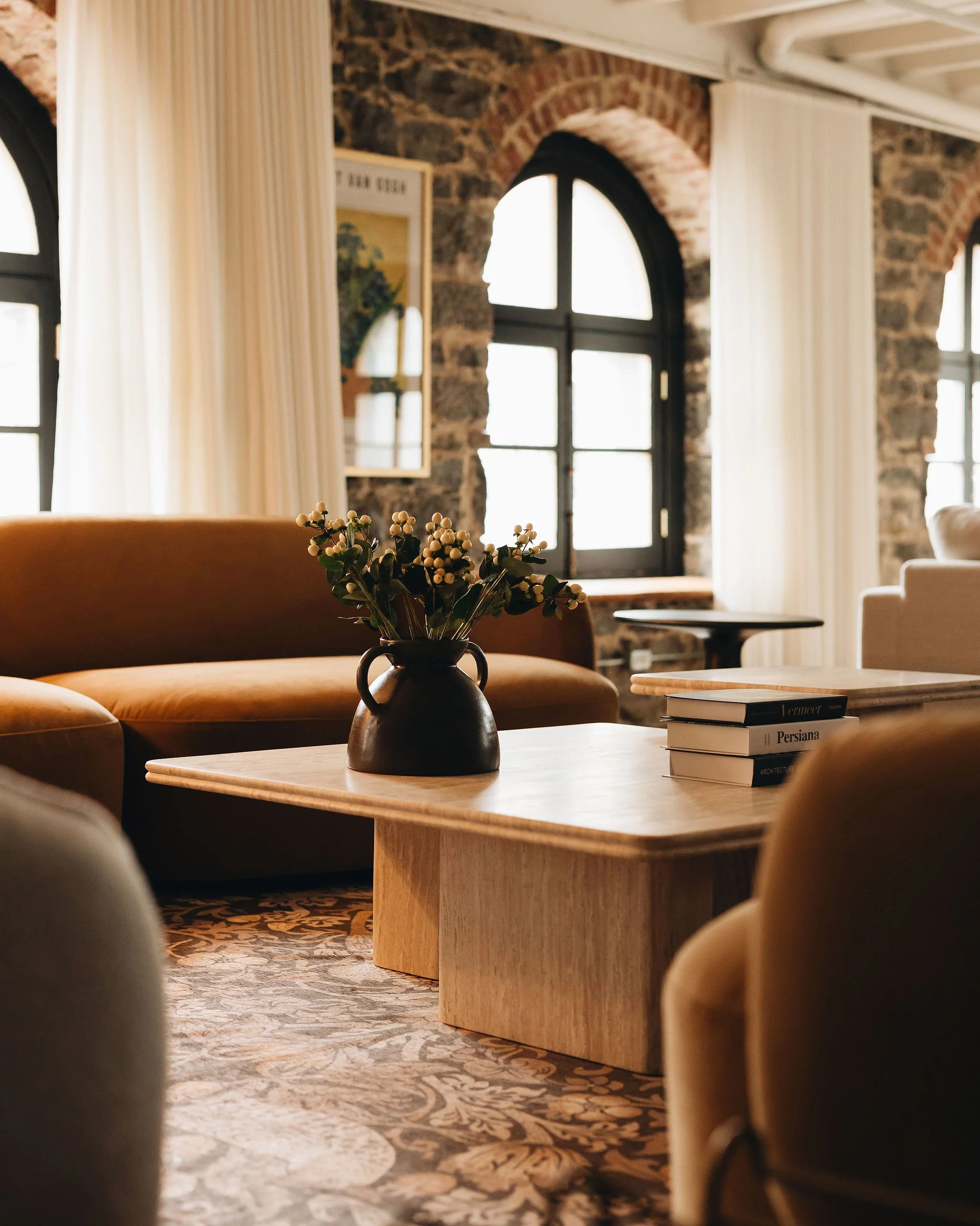 Interior of a room with large arched windows, brick walls, and cream-colored curtains. A wooden table holds a black vase with white flowers, and a stack of books titled 'Persiana' and 'Architecture' is nearby. The room features a patterned carpet and plush furnishings.