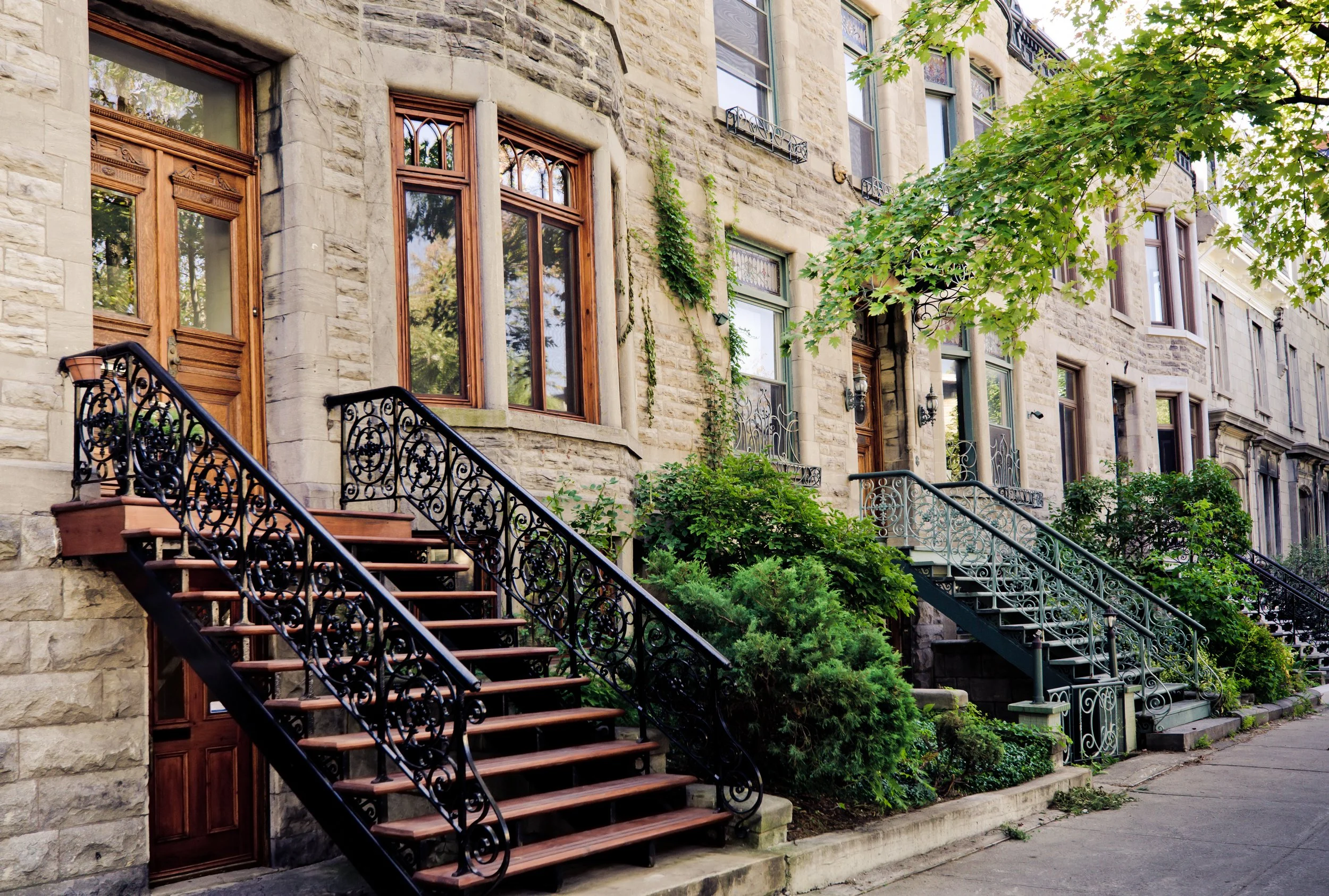 Brownstone building with a staircase leading to wooden doors, surrounded by greenery and multiple windows