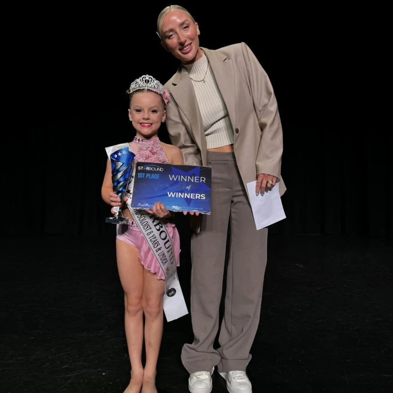 Young Starbound dancer in pink costume with tiara holding a trophy and certificate, standing on stage with special guest adjudicator Claudia Dean.
