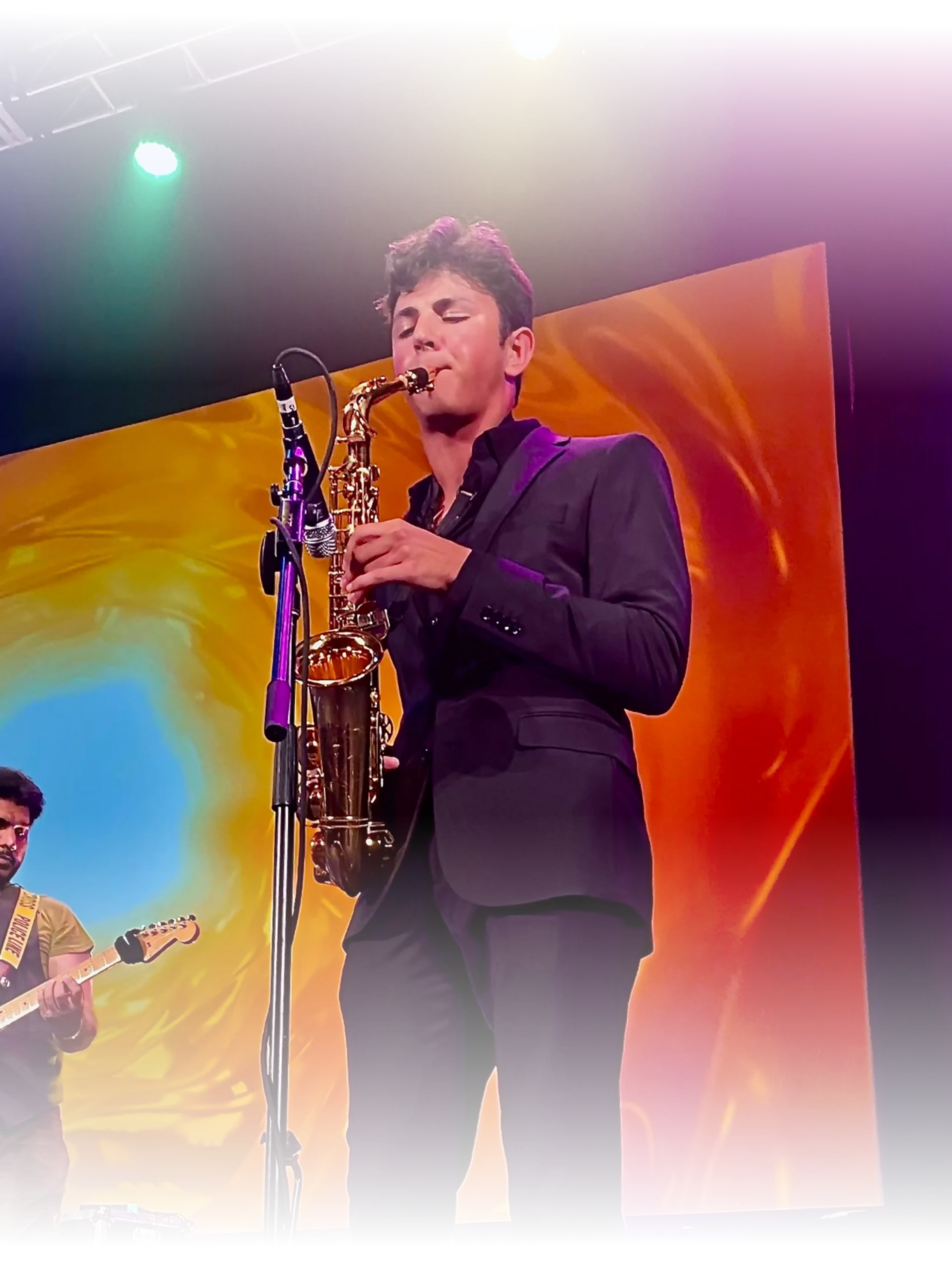A musician in a black suit playing a saxophone on stage with colorful stage lighting and a large abstract backdrop.