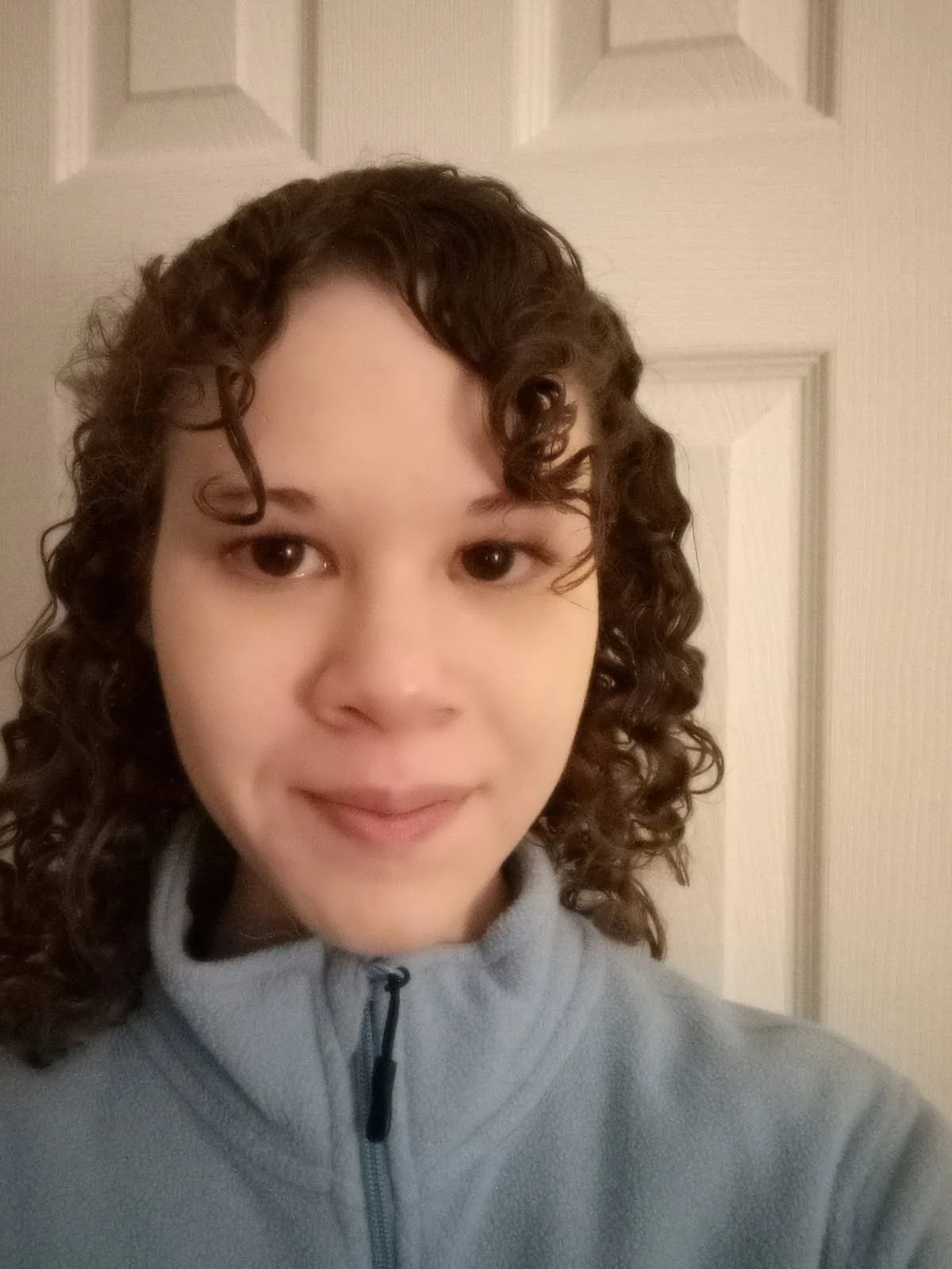 A young woman massage therapist with curly brown hair, wearing a light blue zip-up jacket.