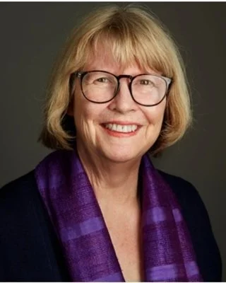 A smiling woman with blonde hair and glasses, wearing a dark blazer and a purple scarf.