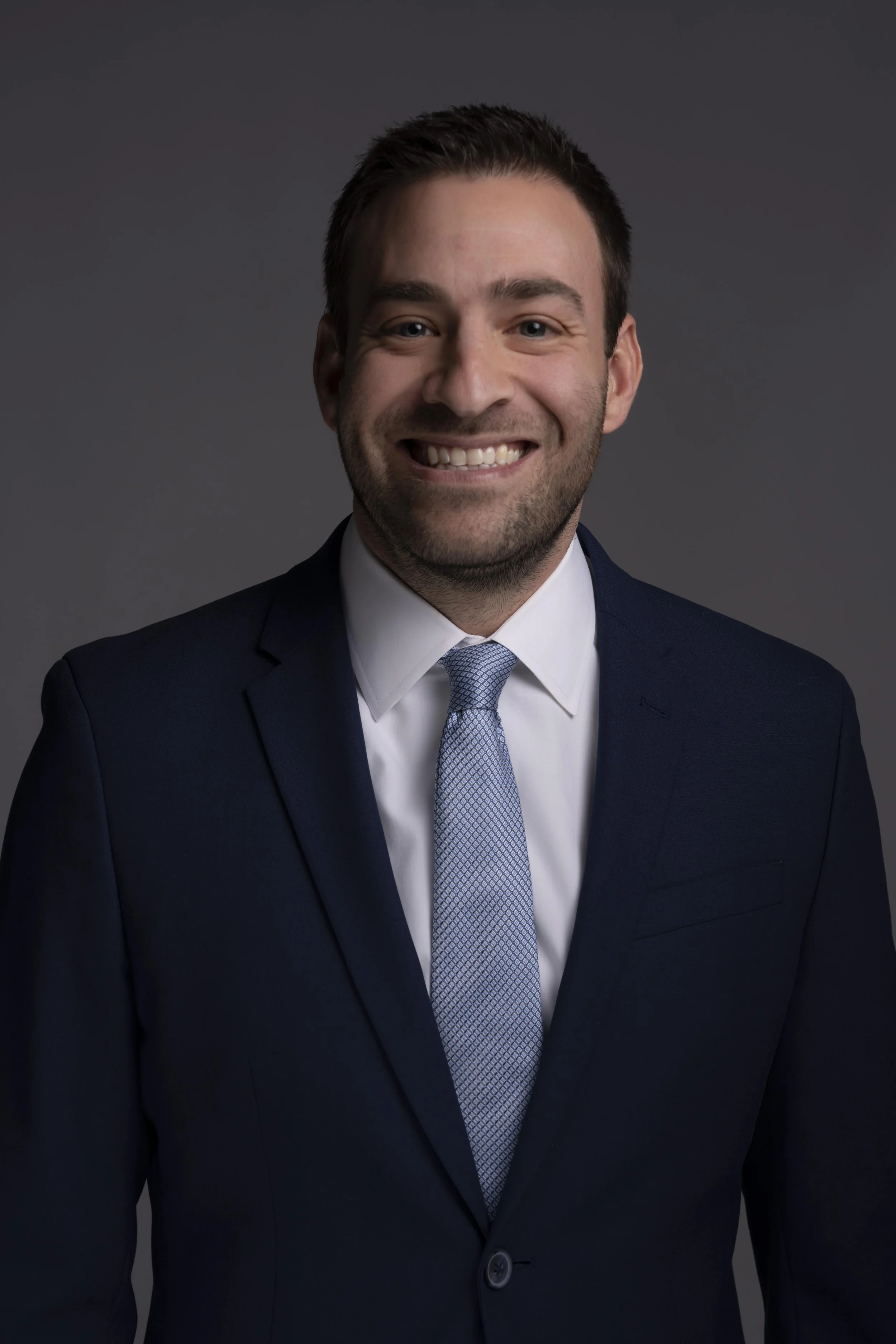 A professional headshot of a smiling man in a navy suit, white shirt, and light blue tie against a dark background.