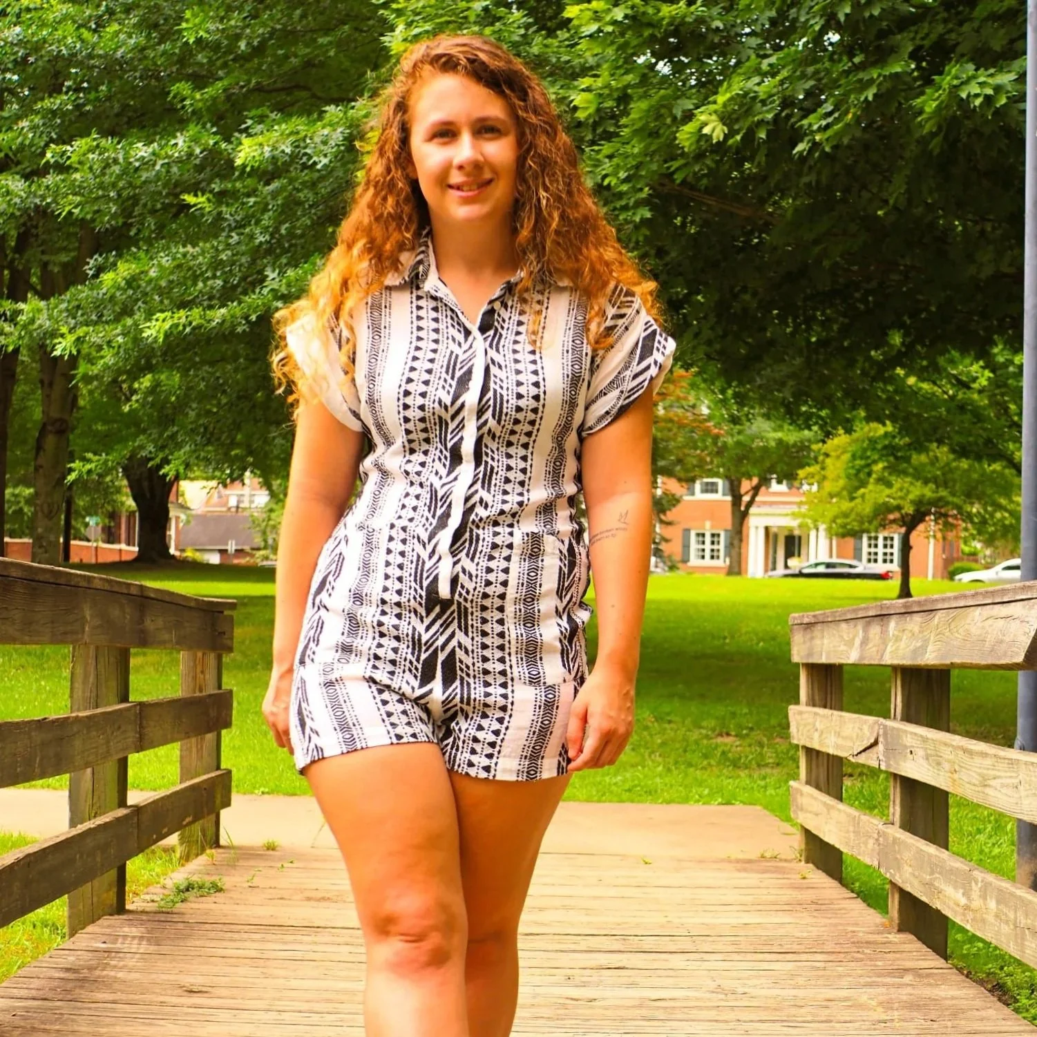 A young woman with curly red hair walks on a wooden bridge in a green park, wearing a black and white patterned romper.