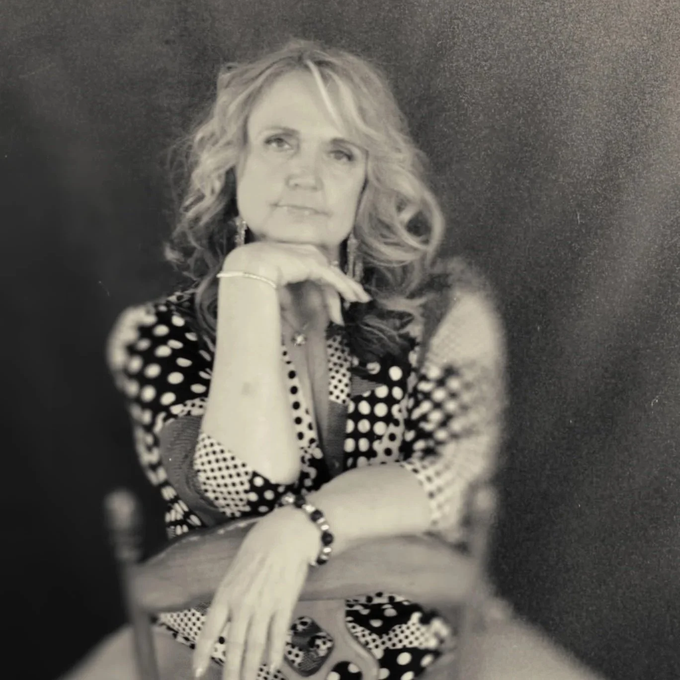 Black and white portrait of a woman with wavy blonde hair, seated with her chin resting on her hand, wearing a polka-dotted dress and accessories.