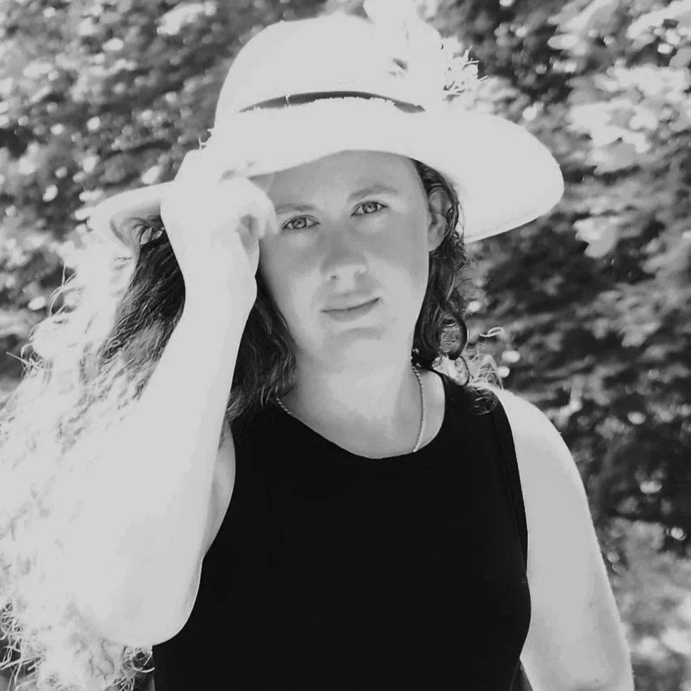 A woman with curly hair wearing a wide-brimmed hat and a black sleeveless top outdoors.