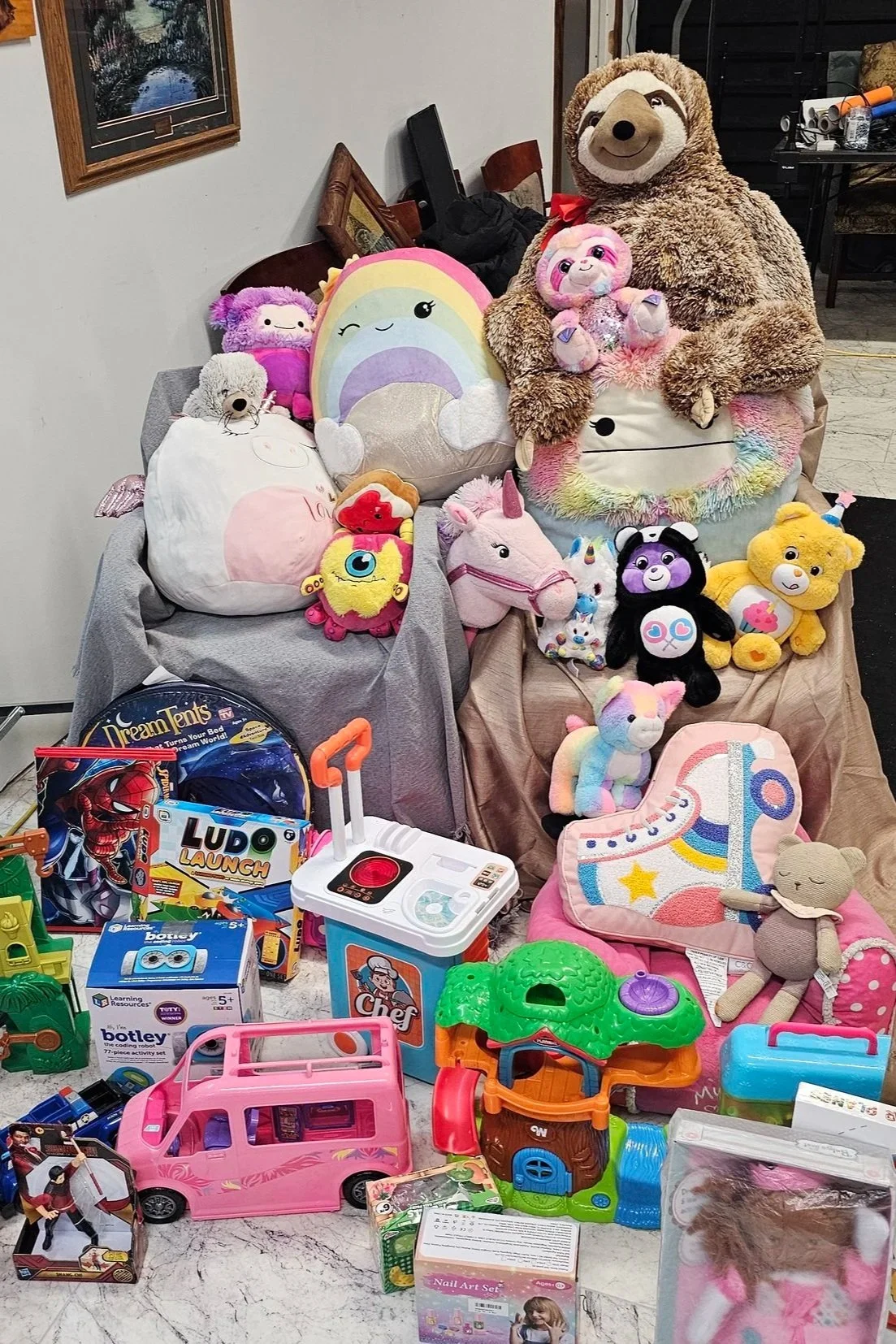 A collection of plush toys, including sloths, unicorns, rainbows, bears, and other stuffed animals, along with various children’s toys such as a pink toy car, a cooking set, and other colorful toys on a table.