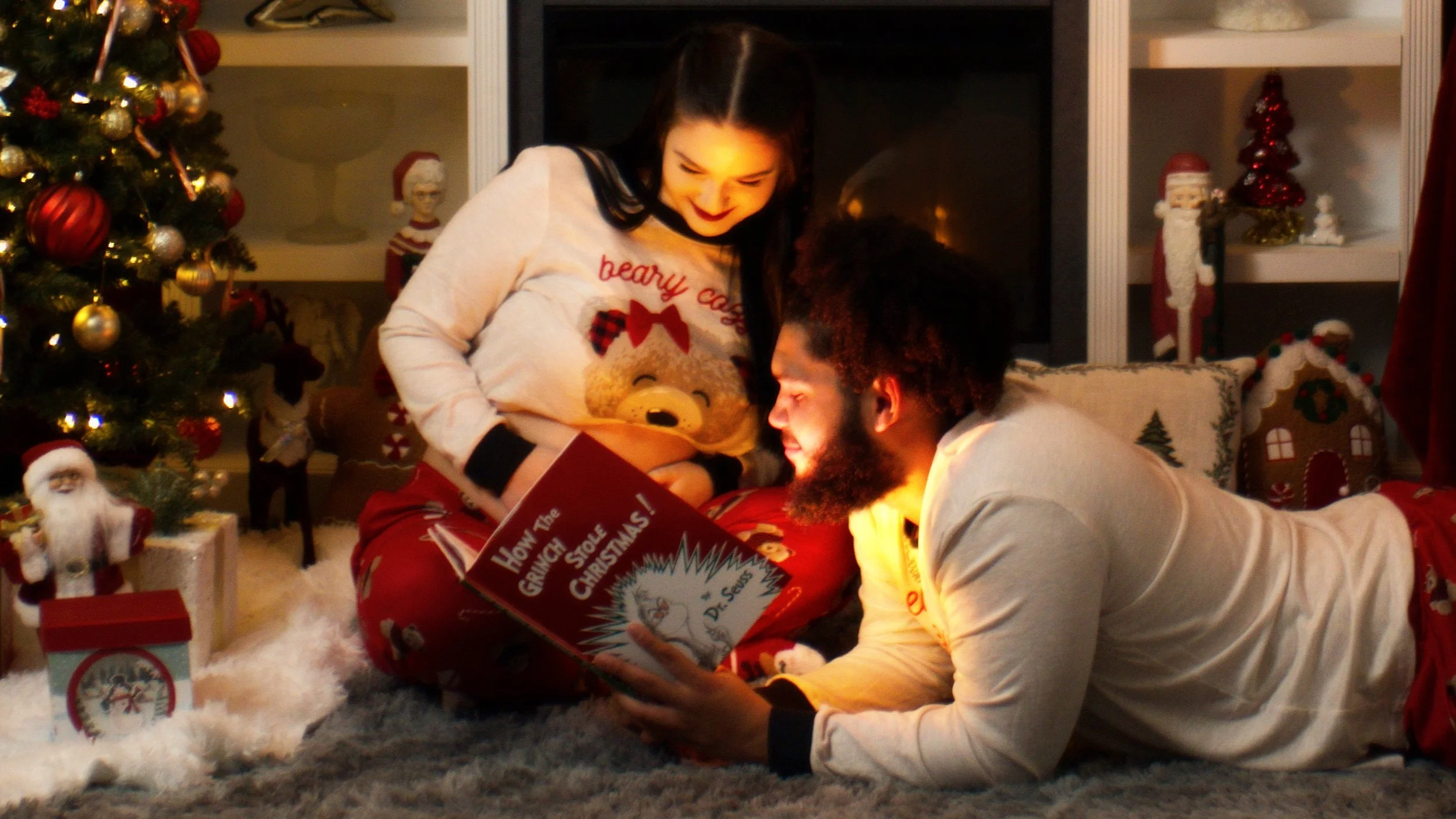 A man and woman in Christmas pajamas look at a book titled 'How The Grinch Stole Christmas' while sitting on the floor in front of a decorated Christmas tree.