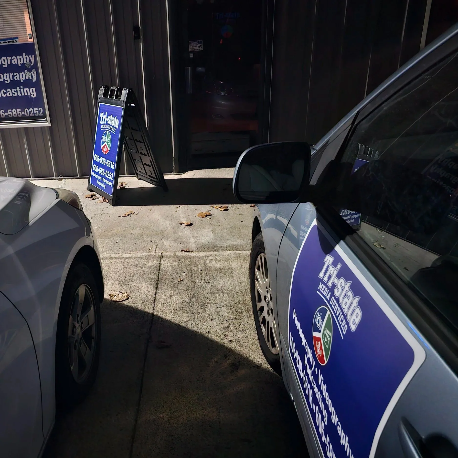 A parking lot outside a media services business with two cars parked, one with signage on the side and another partially visible, and a sidewalk leading to the entrance with a blue signboard displaying contact information.