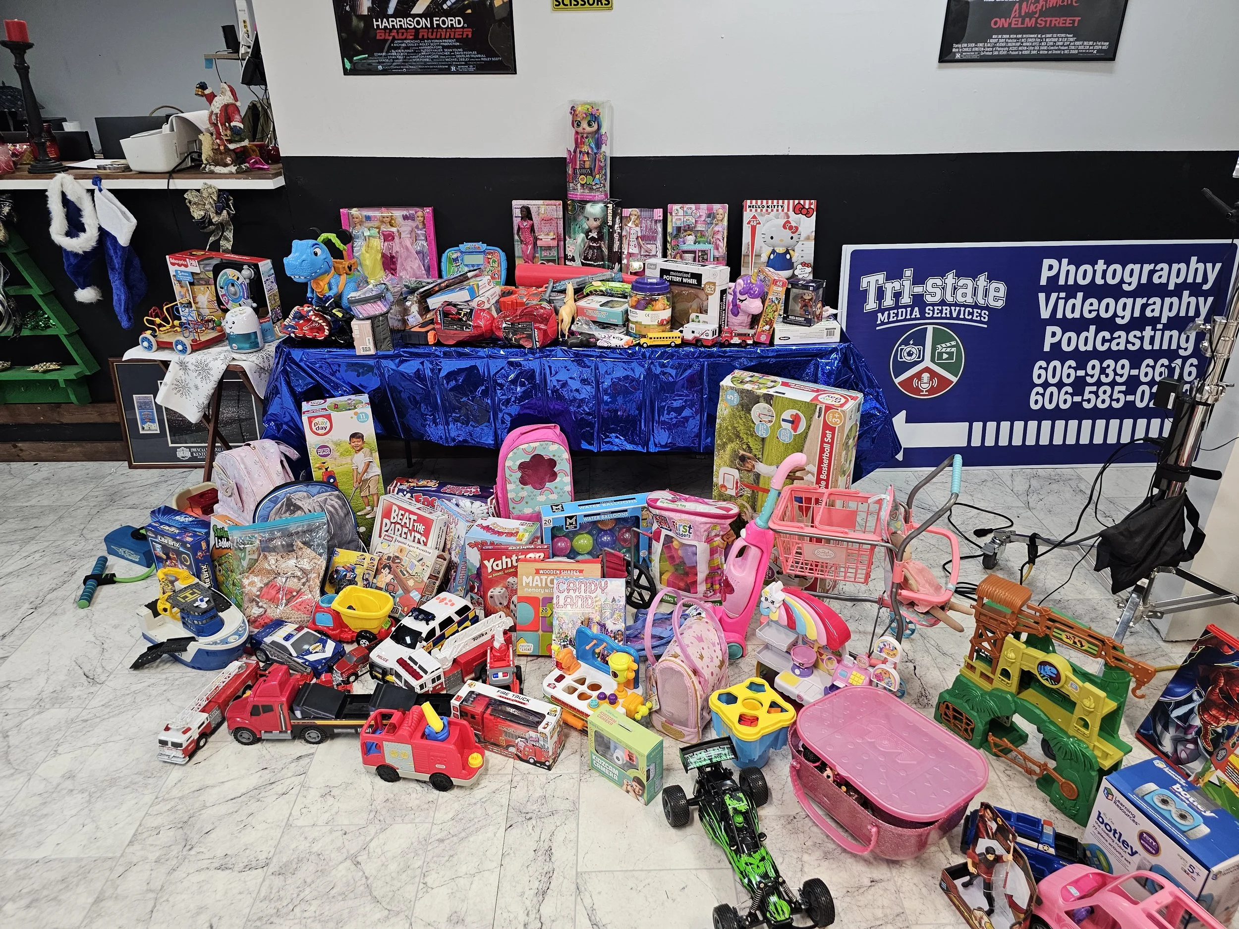 A collection of toys displayed on the floor and on a table at a toy sale or donation event, including stuffed animals, toy vehicles, dolls, and children's accessories. A sign for Tri-state Media Services is visible in the background.