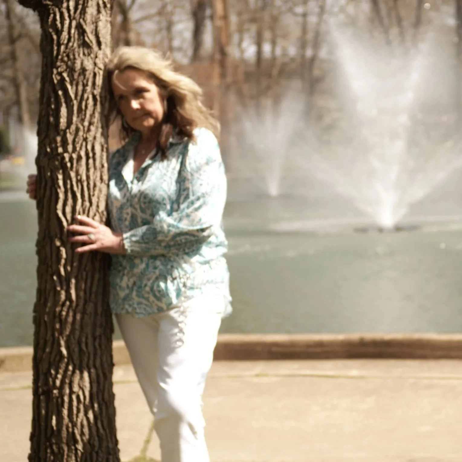 Woman with wavy hair standing behind a tree in front of a fountain in a park.