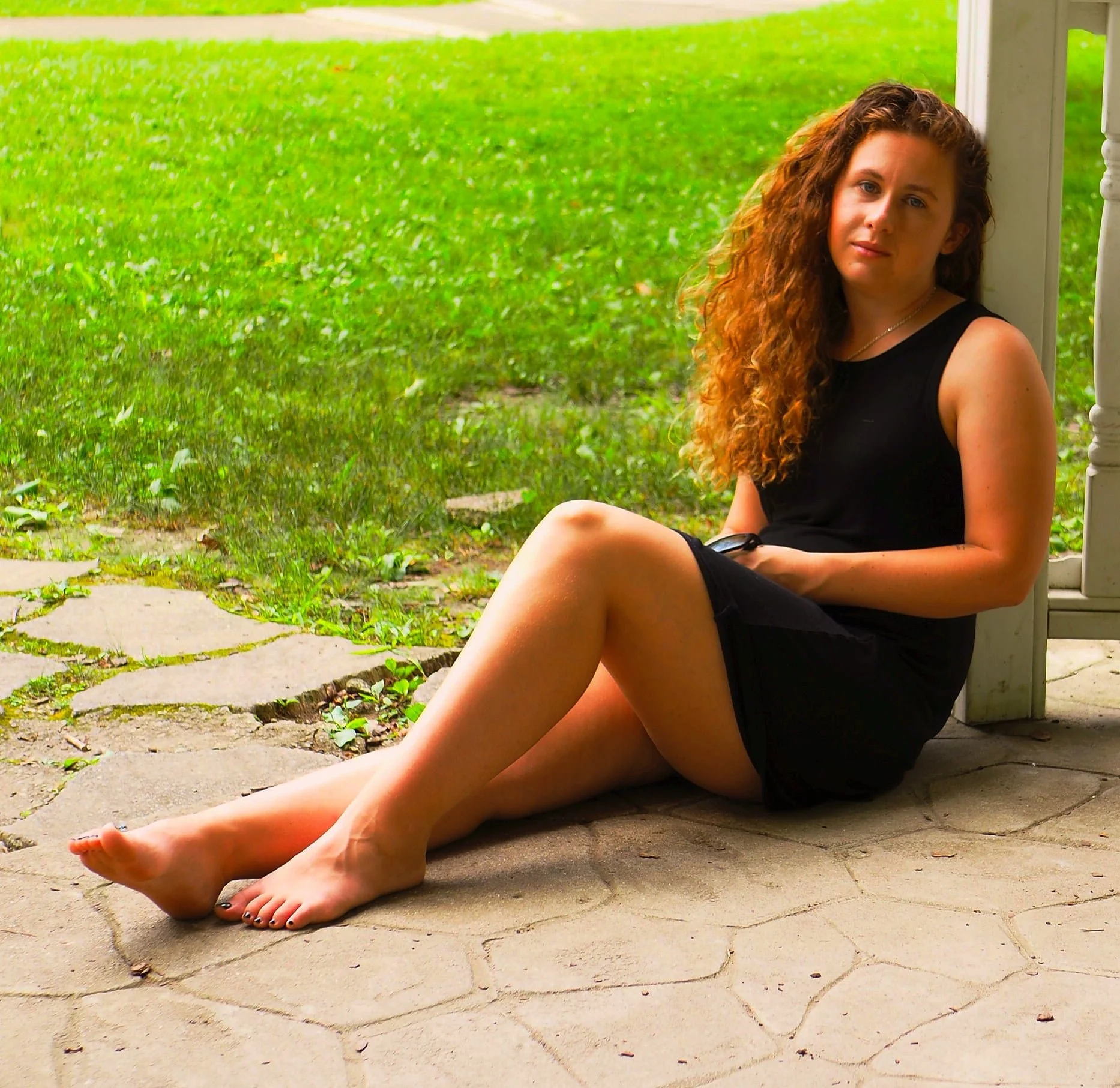 A young woman with curly red hair sits on a stone porch, wearing a black sleeveless top and shorts, holding a remote, with a green lawn in the background.