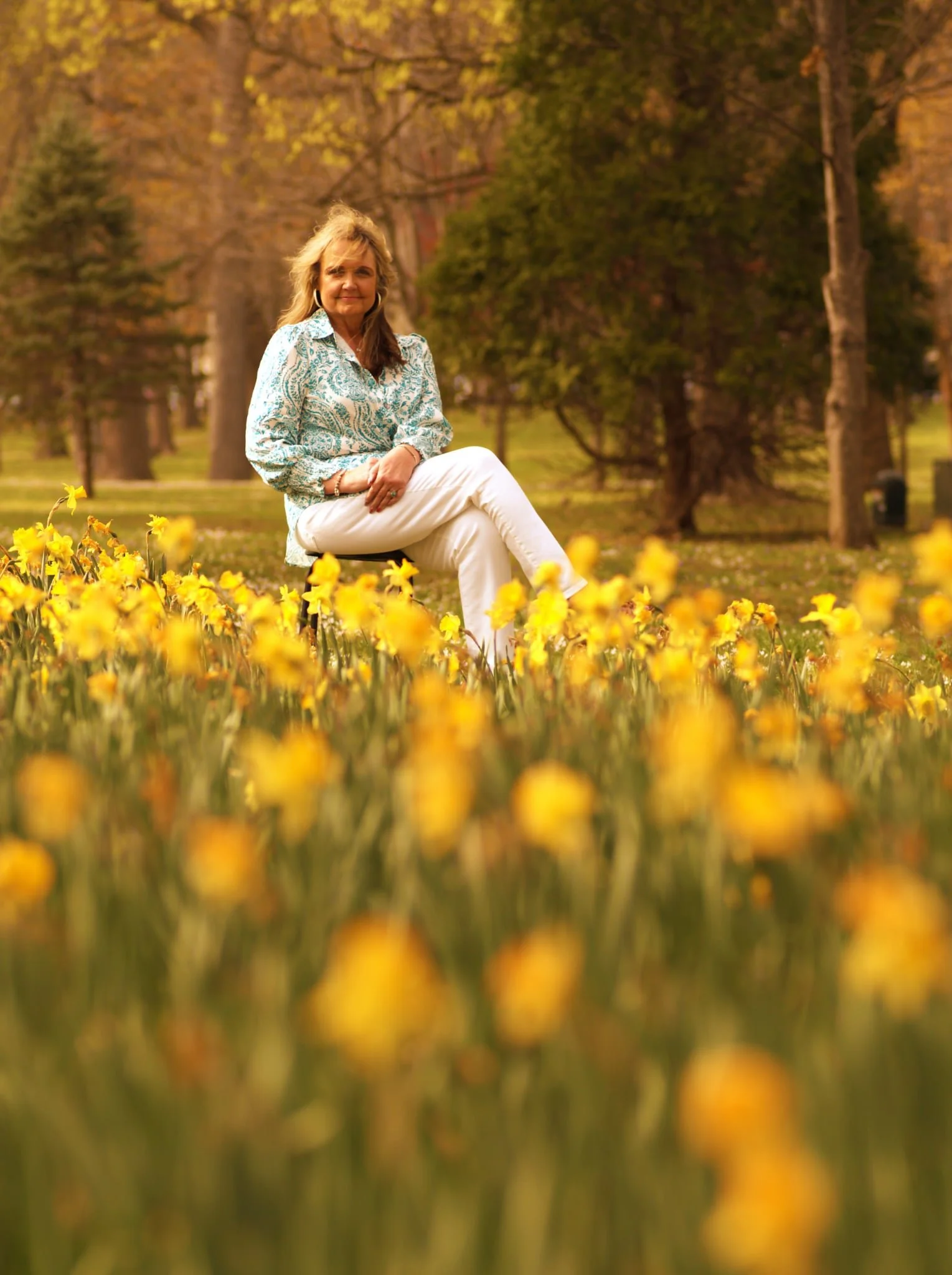 A woman sitting on a bench in a park surrounded by yellow flowers, with trees and fall foliage in the background.