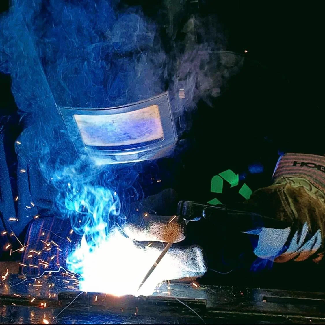 Welding process with sparks and blue light, showing a worker in protective gear and welding helmet.