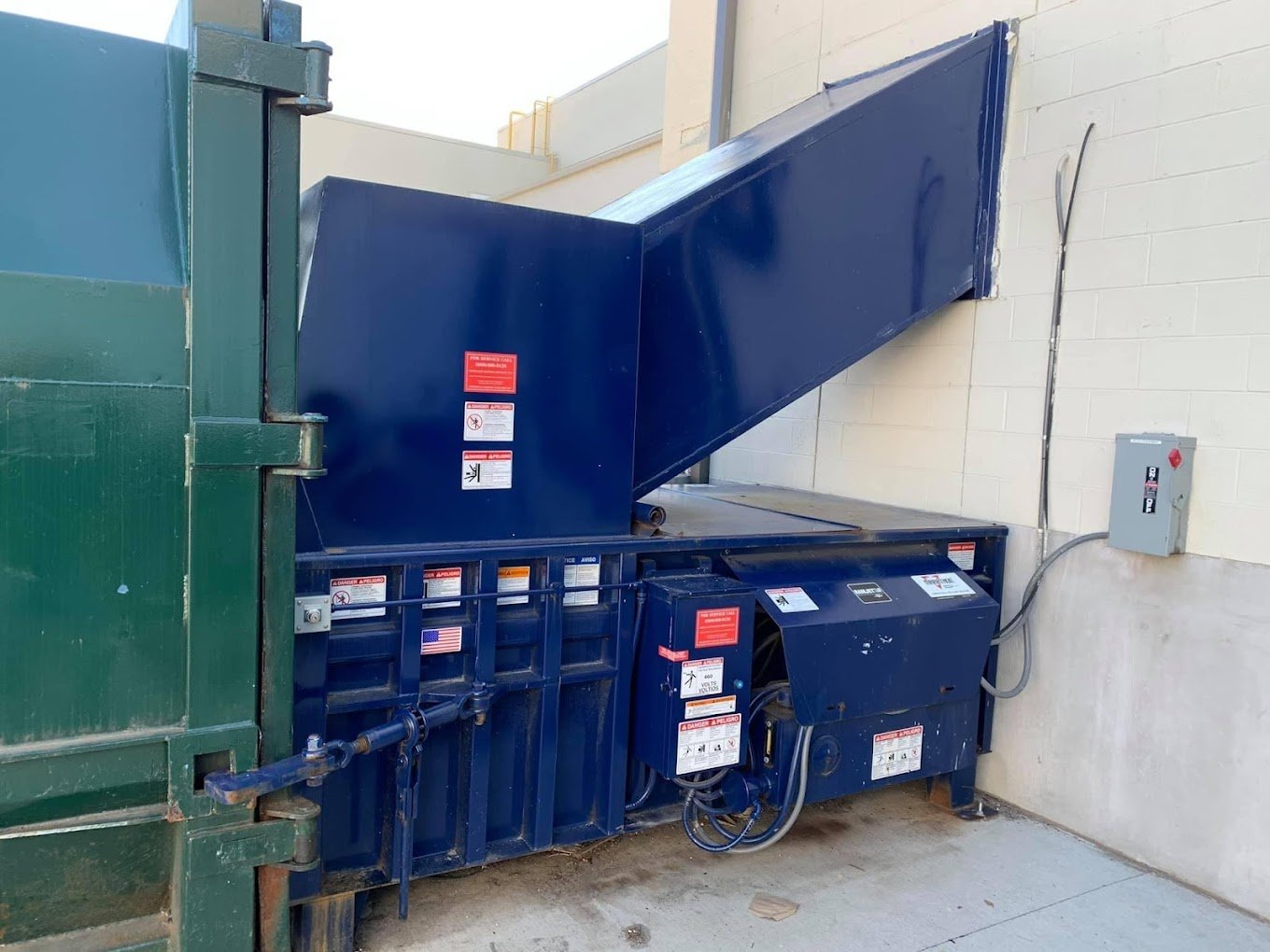 Industrial waste disposal machine outdoors next to a green dumpster, with warning labels and an electrical control box on the wall.