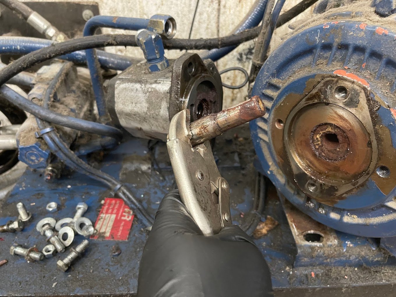 A mechanic's hand holding pliers working on a disassembled blue electric motor or generator with visible rust and metal parts, screws, and bolts scattered on a workbench.